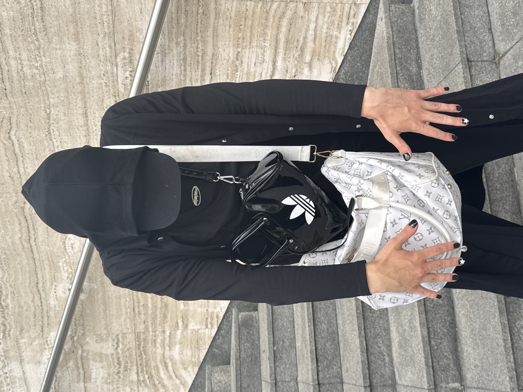 The image shows a person standing on concrete stairs in front of a textured wall. The person is wearing a black hooded jacket, a black t-shirt, a black baseball cap, and black pants. They are carrying two bags: a black patent leather bag with the Adidas logo and a white Louis Vuitton bag. The person's hands are visible, with black nail polish and one fingernail decorated with a white star. The background wall has horizontal striations and a metal railing is visible on the stairs. The lighting suggests an overcast day. This scene is not definitively identifiable as Belgrade, Serbia.