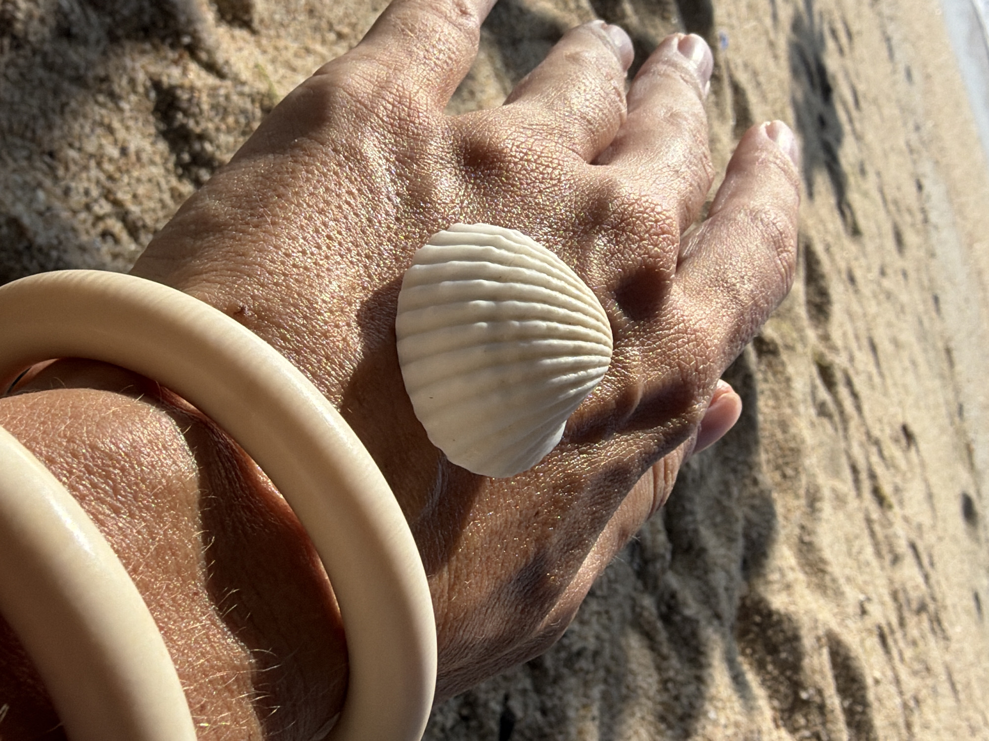 A close-up shot features a person's hand resting on a sandy beach. A small, white, ribbed seashell is placed on the back of the hand. The hand is adorned with a thick, cream-colored bangle. The background consists of the sandy shore with subtle shadows, suggesting it's a sunny day. The image is taken at a low angle, focusing on the hand and the shell, with the beach and a hint of the ocean in the upper left corner. The location is Ko Samui District, Thailand.