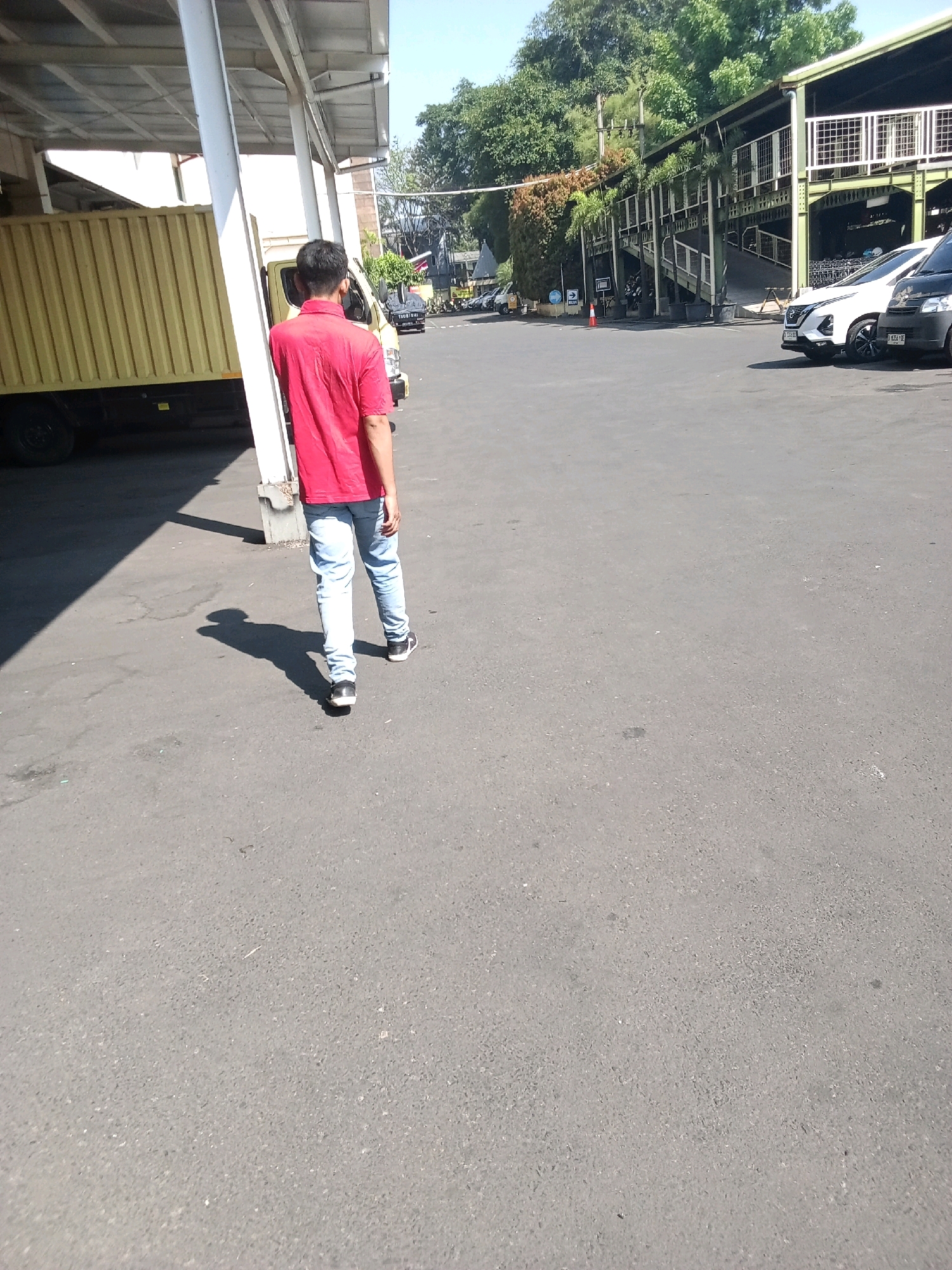 A person walks away from the camera on an asphalt surface, casting a long shadow. The individual wears a red short-sleeved collared shirt and light blue jeans, with dark shoes. To the left of the person is the side of a large yellow truck with a corrugated metal cargo box and a visible black tire. The truck is parked under a white overhang supported by vertical white poles, with a white roof structure above. In the background, a building with a green structure and a white fence extends horizontally. Several cars are parked in front of this building, including a white SUV and a dark-colored vehicle. Trees with lush green foliage are visible behind the building. The scene appears to be an outdoor area, possibly a loading dock or a parking lot, under a clear blue sky. The environment suggests an industrial or commercial setting.