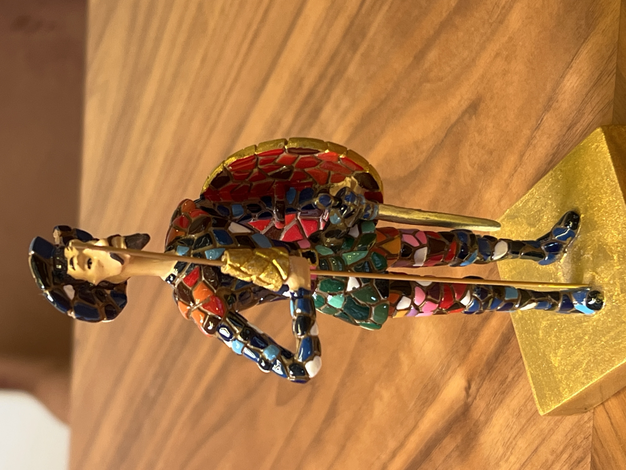 A horizontal, high-angle, close-up shot depicts a colorful mosaic-style statuette of a matador resting on a light brown wooden surface, with a bright gold, pyramid-shaped base visible at the bottom right. The matador figure is dressed in a multi-colored outfit composed of small, irregularly shaped tesserae in shades of red, blue, green, orange, pink, and white, with gold accents. He has a fair-skinned face and wears a dark blue hat. The figure holds a long, thin golden object across its body. The location of Yerevan, Armenia, is not verifiable.