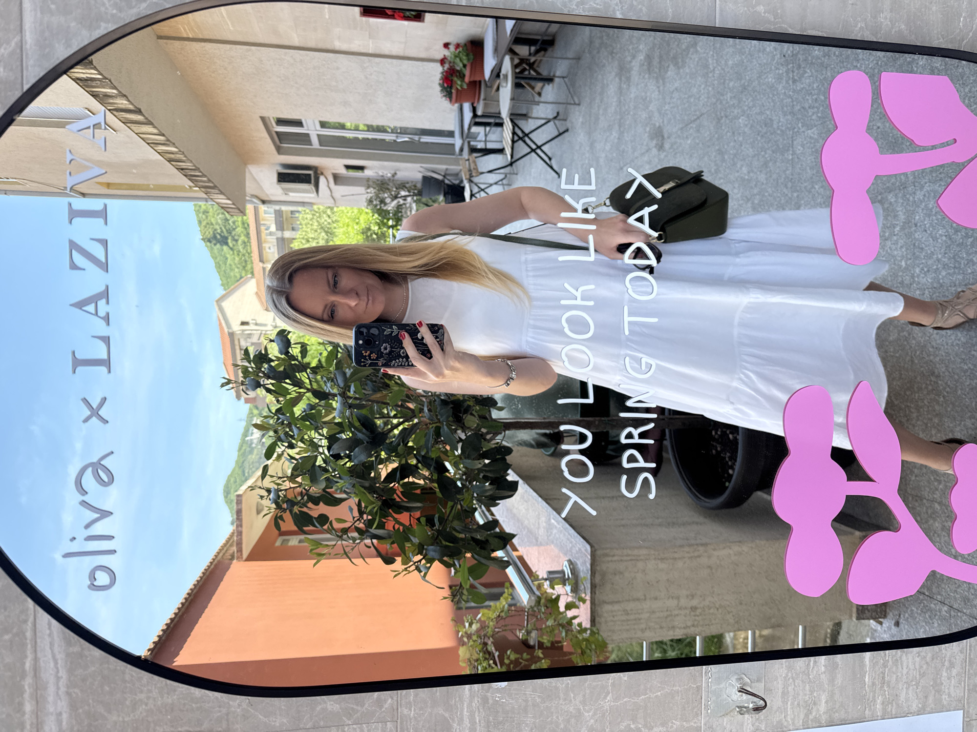 A woman in a white dress is taking a selfie in a mirror that has "oliva x LAZIZA" written on the top left corner, "YOU LOOK LIKE SPRING TODAY" written in the middle, and pink flower-like decorations on the right. The mirror reflects an outdoor setting, with buildings and foliage visible in the background. The image likely originates from Budva, Montenegro, based on the "LAZIZA" text, which is associated with a location in Budva. A small green purse hangs on the woman's shoulder. She is also holding sunglasses. The setting appears to be a patio or outdoor cafe, with tables and chairs seen in the mirror's reflection.