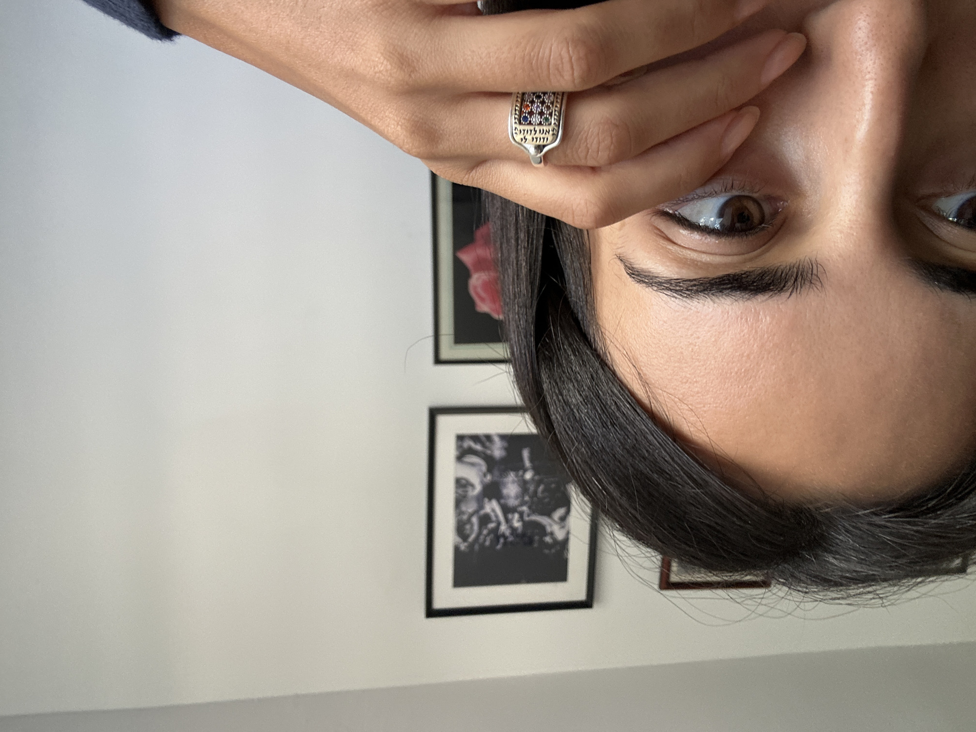 A close-up, eye-level shot shows the left side of a person's face, focusing on their eye and forehead. Their dark hair frames the face, and their left eye, with brown irises and dark lashes, is visible and looking slightly to the left. A hand with painted fingernails is raised, with fingers gently touching the cheek and near the eye. A ring with colorful gemstones and inscribed text is worn on the ring finger. In the background, a white wall is adorned with two framed pictures. The top frame contains a black and white image of people in motion, possibly dancing. The bottom frame displays a close-up of a pink flower. The lighting suggests an indoor setting with natural light coming from the right. The scene is devoid of any explicit Tel Aviv landmarks, making its specific location within the city indeterminate based solely on the visual information.