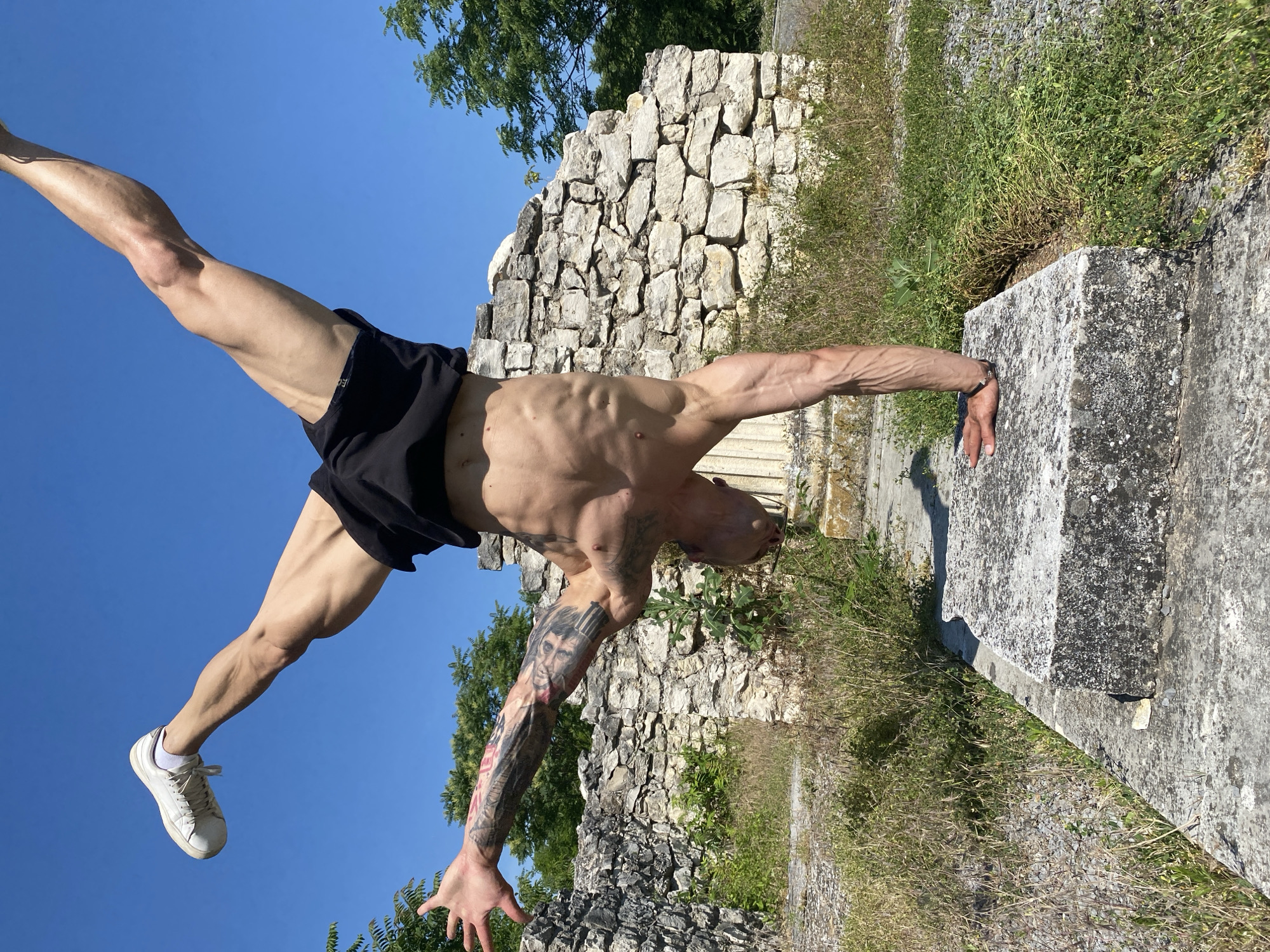 A muscular, shirtless man is performing a handstand on a stone block in front of a stone wall ruin. He is wearing black shorts and white sneakers. His legs are spread wide apart in a V-shape, and his left arm is extended upwards. The right arm is bent at the elbow and supporting his weight. The man has tattoos on his left arm and chest. The background consists of the stone wall, overgrown vegetation, and a clear blue sky. The scene appears to be outdoors in a natural or historical setting. This location is the St. George's Chapel ruins in Pleven, Bulgaria.