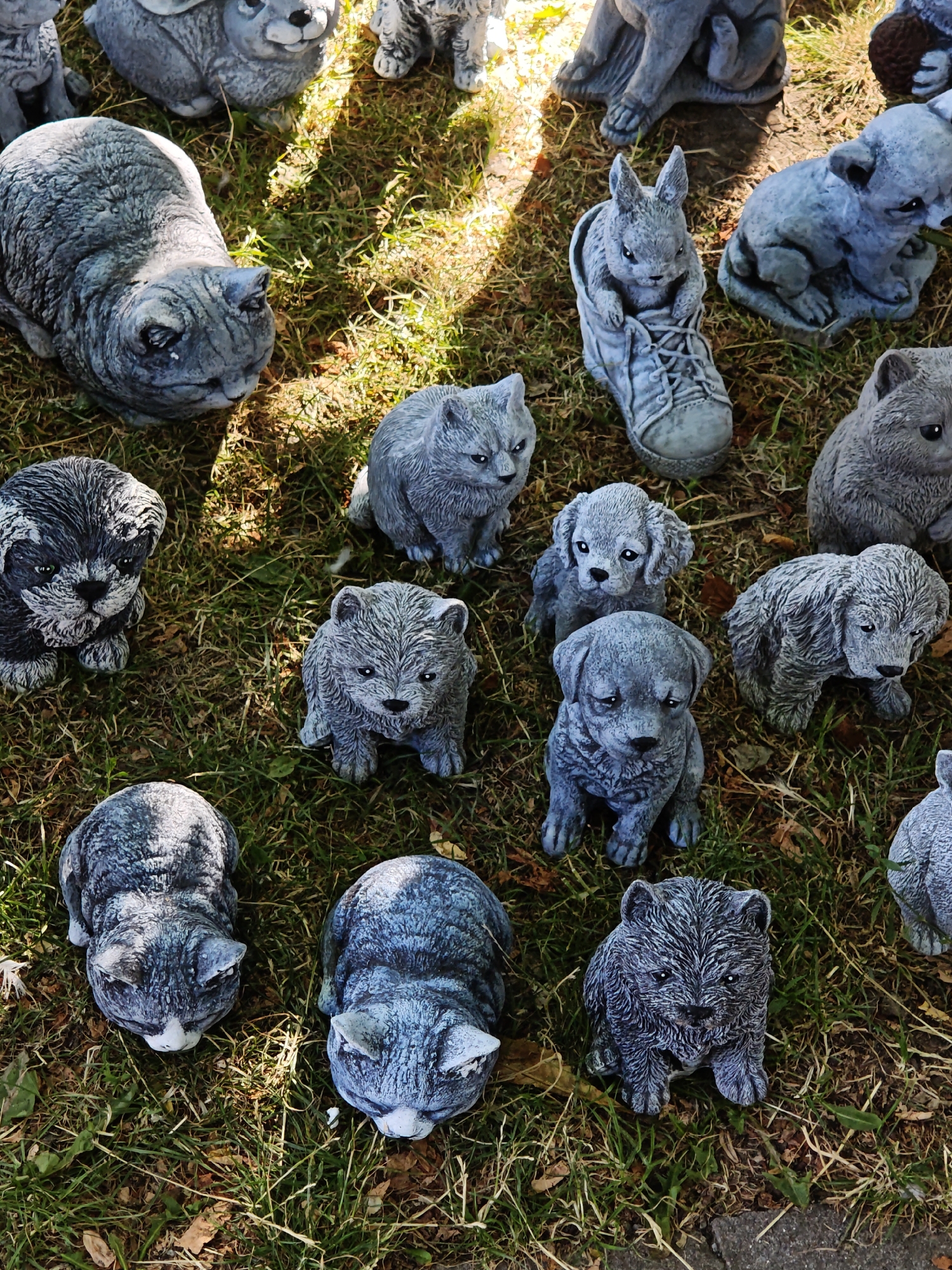 A collection of gray, stone animal figurines is displayed outdoors on a patch of grass and dirt, suggesting a garden or outdoor market setting. The figurines include several cats, dogs, and what appears to be a rabbit in a shoe. The scene is illuminated by sunlight, creating shadows on the ground. The location is identified as Vinnytsia, Ukraine.
