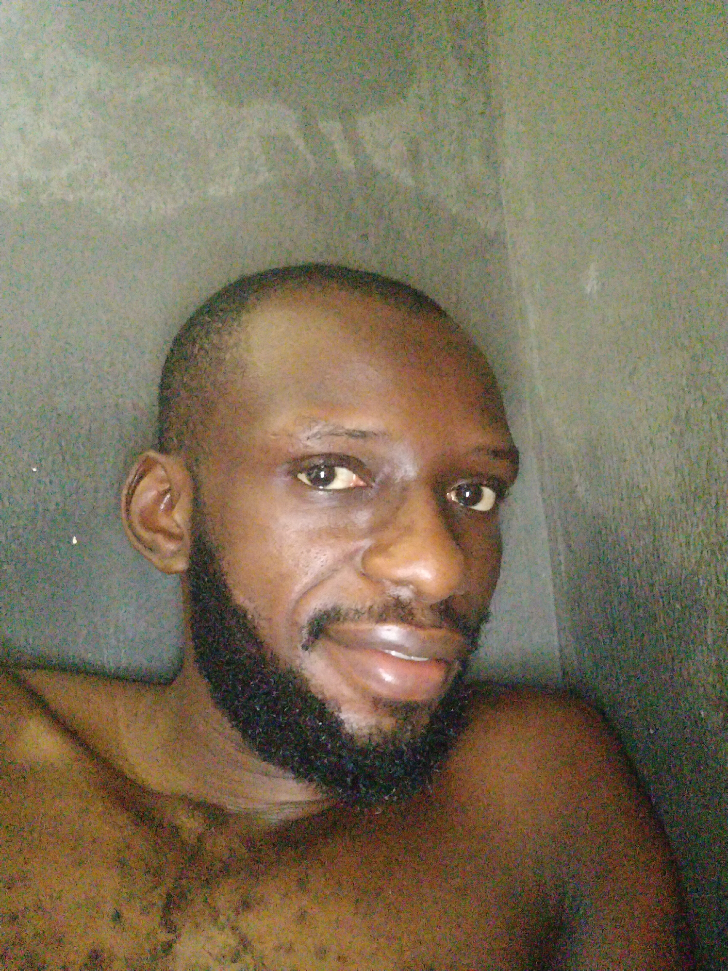 A close-up portrait of a dark-skinned man with a shaved head and a full beard. The man is positioned between two gray walls. The location is likely indoors, potentially in Uyo, Nigeria.