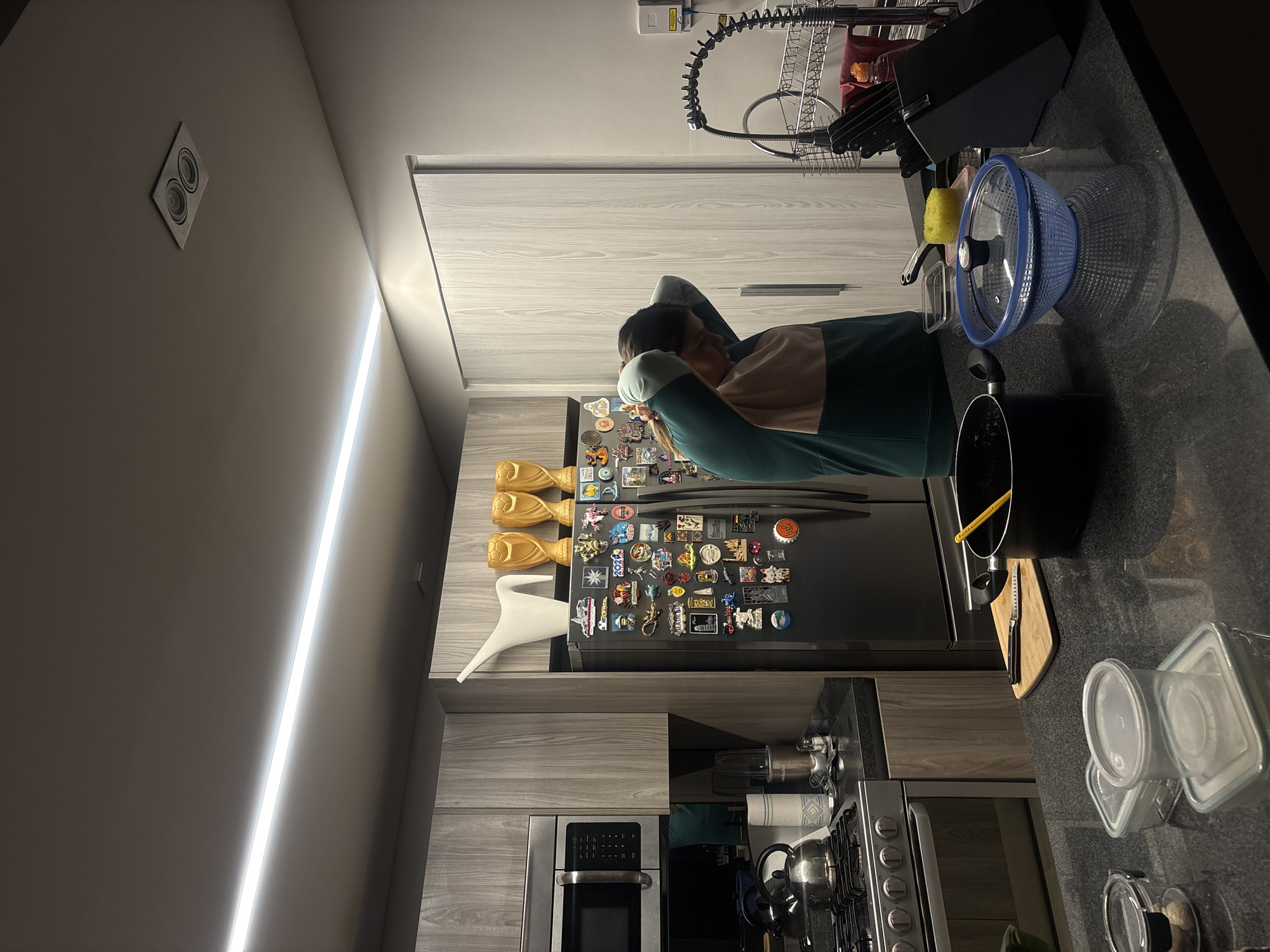 The image shows a kitchen interior, likely a residential space. A person is leaning against a refrigerator with their arms crossed behind their head. The refrigerator is covered in magnets. Countertops are visible on the right, with a black pot, a blue colander, containers, and kitchenware. A stove and oven are below the counter. The walls and cabinets are a light gray wood grain. A long, linear light fixture illuminates the scene. A dish rack hangs above the counter. The exact address in San Bernardino Tlaxcalancingo, Mexico is not possible to determine from this image.