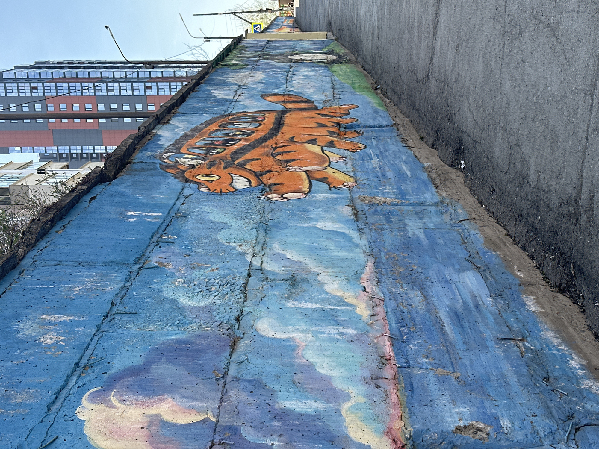 A low-angle shot captures a concrete retaining wall in Novosibirsk, Russia, adorned with street art. The wall is painted blue, featuring cloud-like patterns in the lower portion and a large, cartoonish turtle-like creature painted in orange with black stripes and visible teeth near the top. The mural extends along the wall. A tall building with multiple windows is visible in the background on the left, partially obscured by vegetation near the base of the wall. The top of the wall runs uphill, with a gray concrete wall visible to the right. Street signs can be seen further up the slope.