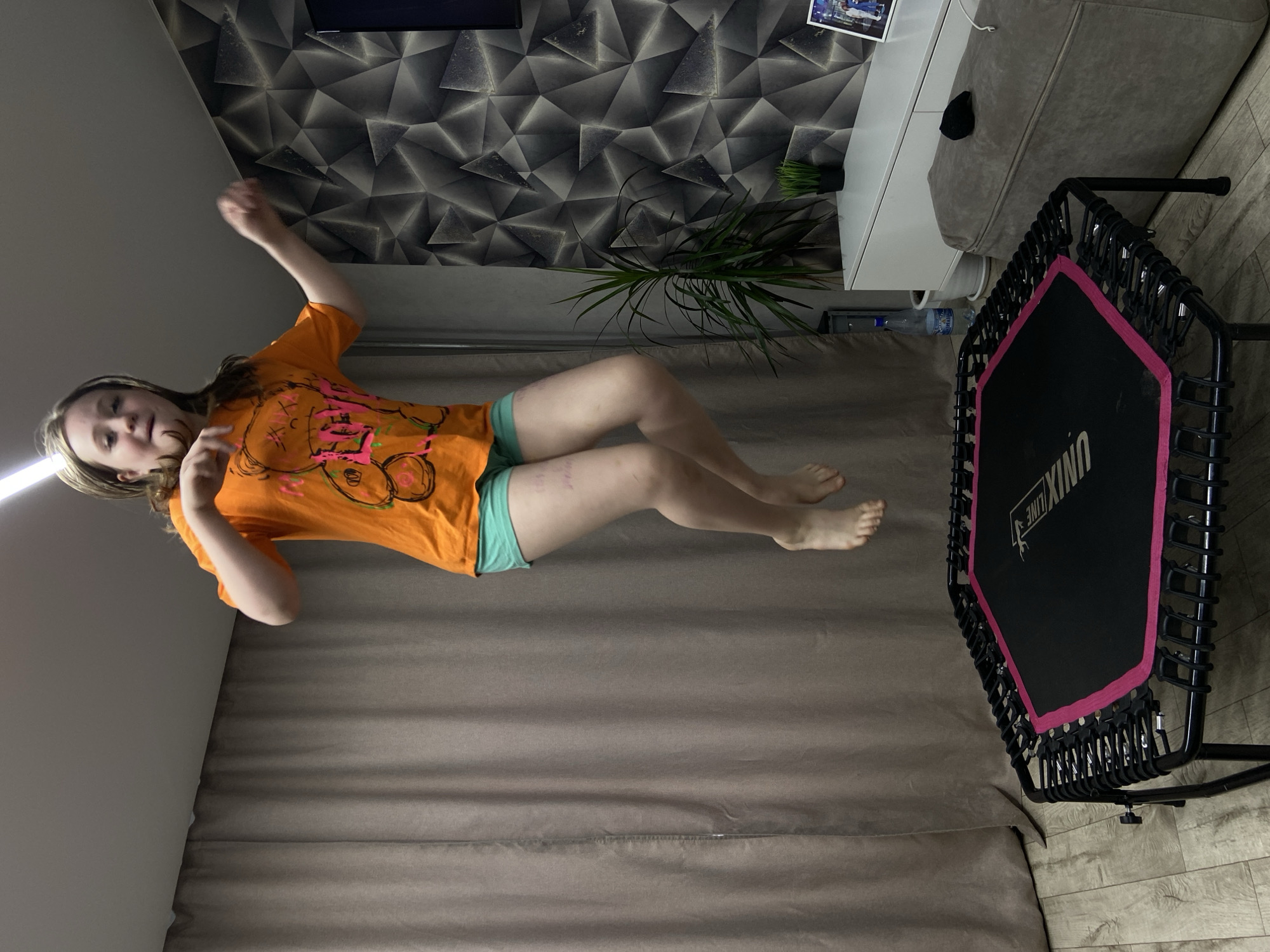 A young girl is captured mid-air, seemingly jumping on a mini-trampoline, within an indoor room in Chaikovsky, Russia. The image is oriented sideways, giving the impression of her floating horizontally. The girl has light brown hair and is wearing an orange t-shirt with a colorful graphic design on the front, and light blue-green shorts. Her arms are raised and bent, and her legs are drawn up, indicating she is in motion. Her expression appears focused or mildly pleased.
The black mini-trampoline with pink trim is positioned to the right of the girl, featuring the brand "UNIXLINE" prominently on its mat. The room's background includes a wall covered in a dark, geometric patterned wallpaper, behind which a green houseplant in a black pot is visible. To the right, a white low cabinet or console table holds a picture frame and other small objects. A grey upholstered sofa is partially visible behind the trampoline and cabinet. The floor is covered with wood-look laminate, and a brown curtain or blind extends across the lower portion of the wall. A plastic water bottle with partially legible Russian text, "ШИШ", is on the floor near the sofa. The lighting suggests an indoor setting, likely relying on artificial light.