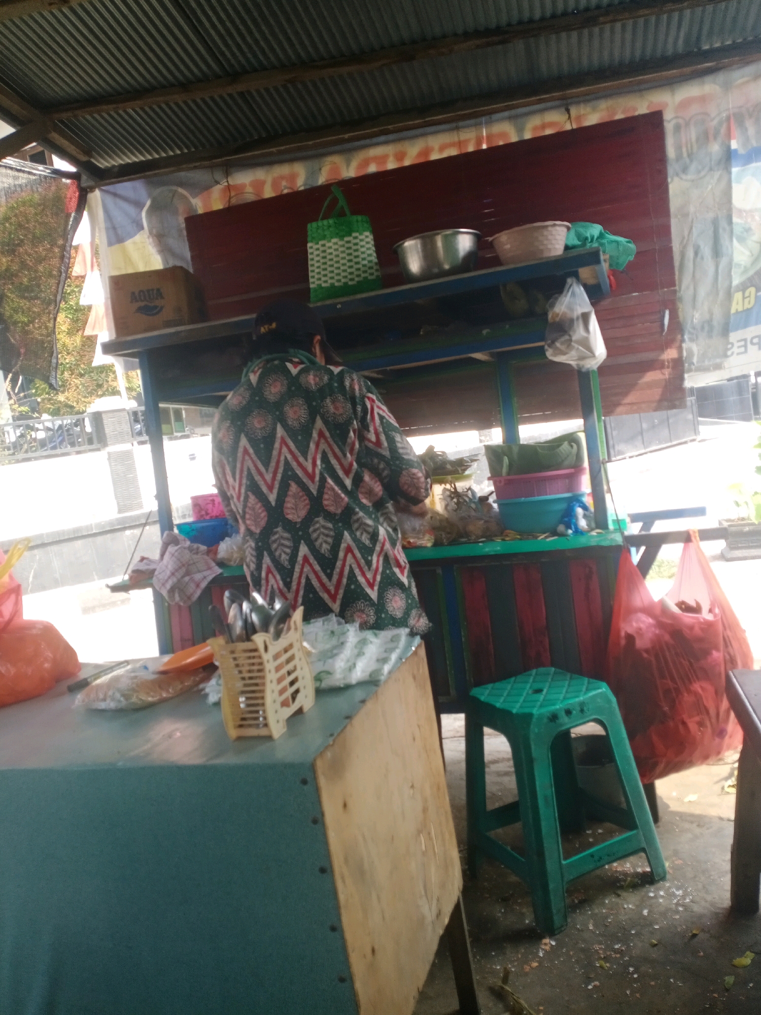 A woman wearing a patterned shirt stands behind a food stall in Samarinda, Indonesia. The stall has a corrugated metal roof and is decorated with colorful stripes. On a shelf above the counter, there are bowls, baskets, and a green woven bag. In front of the woman, on the counter, are various items including a plastic bag, a yellow organizer with utensils, and an orange plastic bag. A green plastic stool is positioned to the right of the stall. The ground around the stall is paved and scattered with debris. A large red bag is visible to the right of the stool.