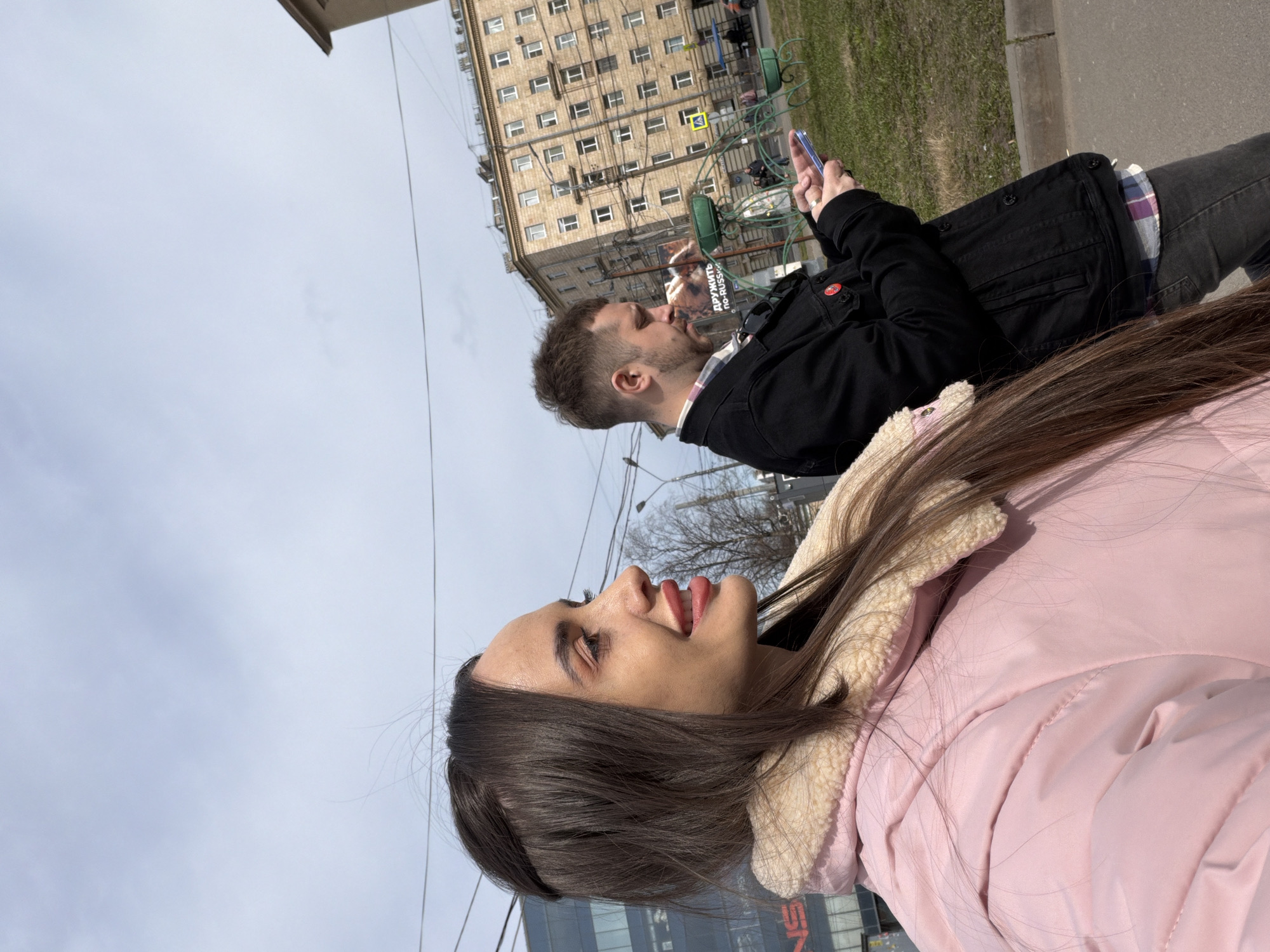The photograph shows two individuals, a woman and a man, seated outdoors in an urban setting, with a building in the backdrop. The woman is in the foreground, wearing a pink padded jacket with a cream-colored fleece collar. Her brown hair falls over her shoulder. She appears to be looking and smiling upwards. Behind her, a man is seated, wearing a black jacket over a plaid shirt and dark jeans. He holds a mobile phone in his hand, which he appears to be looking at.
In the background, a multi-story building with multiple windows and architectural details is visible, indicating an urban area. A pedestrian crossing sign is visible next to the building. Street lights and overhead wires can also be seen. Given the presence of Russian writing on a poster in the distance, the photograph is likely taken in Russia, specifically in Saint Petersburg.