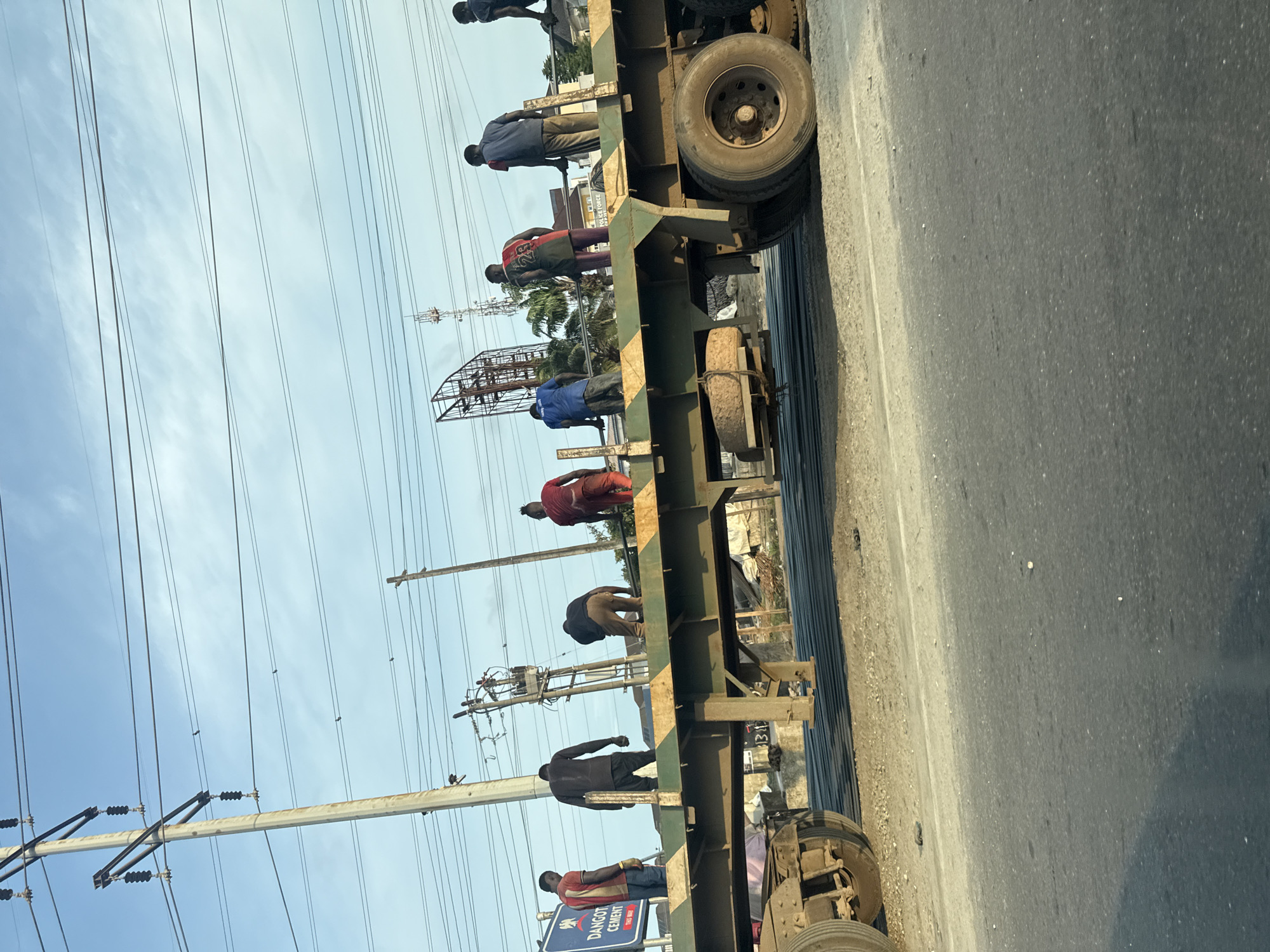 In Lagos, Nigeria, several individuals are standing on the flatbed of a large truck. The truck appears to be carrying metal rods, which are visible stacked on the trailer. The people are diverse in their attire, wearing a mix of casual clothing such as t-shirts and pants, some with visible numbers on their shirts. Their postures suggest they are either working or observing. The environment shows a road with a sandy shoulder and a clear sky above, dotted with utility poles and power lines, indicating an urban or semi-urban setting. A sign for "Dangote Cement" is visible, suggesting the location is within the industrial or commercial areas of Lagos where such businesses operate. The scene is captured from a slightly elevated angle, likely from a vehicle passing by.