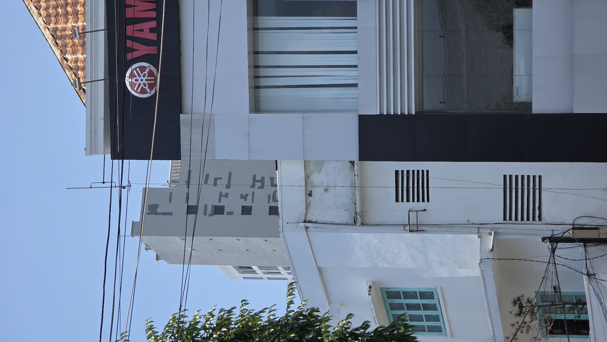 The image shows a vertical view of a building in Nha Trang, Vietnam, under a clear blue sky. The building displays a large "YAMAHA" sign, positioned next to windows with white vertical slats and a dark reflective window. Below, there is a lighter section of the building with square ventilation grills, light-blue windows, and power lines are visible in the foreground, along with a tree. Another building is seen behind, with a pattern of square windows.