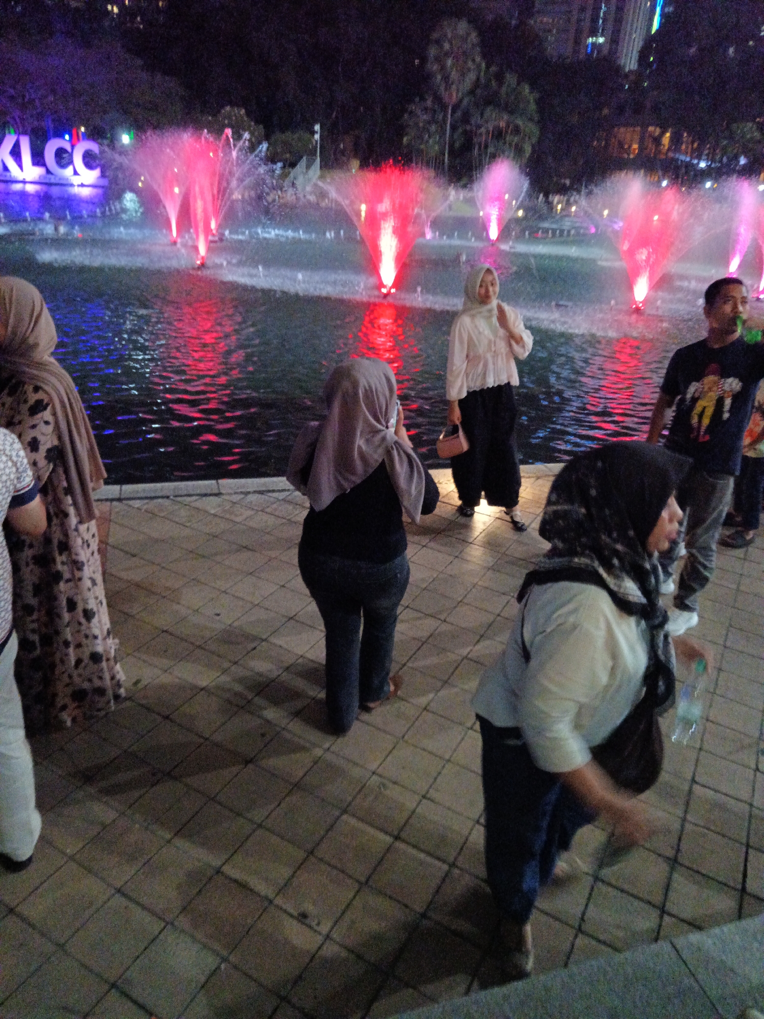 The image depicts an outdoor scene at night in Kuala Lumpur, Malaysia, likely near the Petronas Twin Towers, as suggested by the "KLCC" illuminated sign in the background. A large fountain is visible with vibrant red and pink water jets, reflecting on the water's surface. Several individuals are present in the foreground, some appearing to be tourists or locals enjoying the attraction. One woman, wearing a light-colored headscarf and a patterned dress, is seen from the side. Another person in the foreground is wearing a grey headscarf and a black shirt. A man in a black t-shirt with a graphic print is standing to the right, holding a green bottle. The ground is paved with square tiles, and the overall atmosphere suggests a lively public space with illuminated attractions.