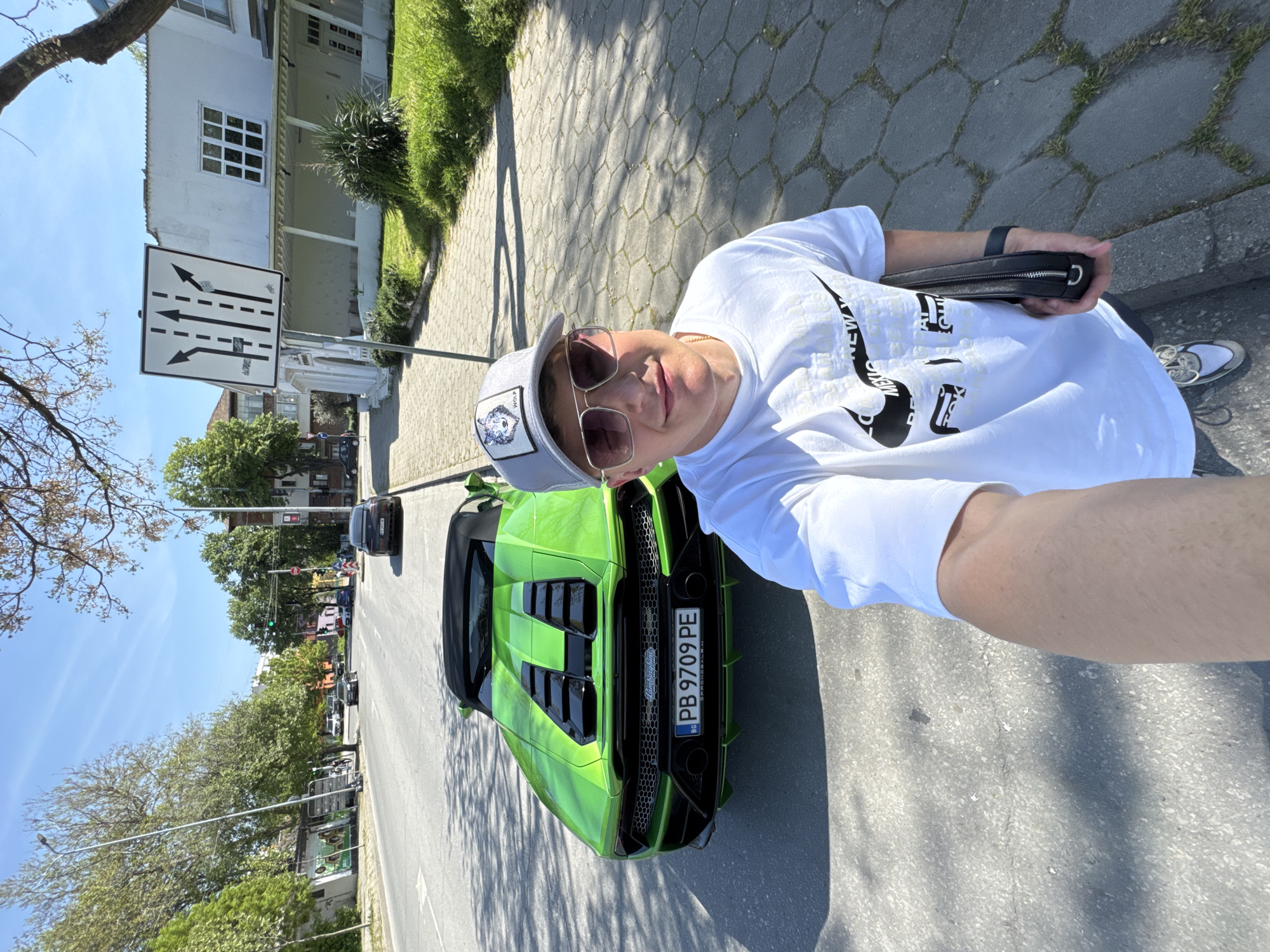 A selfie image, captured on a bright, sunny day in Plovdiv, Bulgaria, depicts a person posing next to a vibrant lime green Lamborghini convertible. The photo is tilted, presenting a dynamic view of the street scene.
The primary subject is a light-skinned individual, seen from the chest up, wearing a white short-sleeved t-shirt with a black graphic print (partially visible "NEW YORK"), square-framed sunglasses, and a grey baseball cap adorned with a wolf logo patch. They are holding a small black clutch bag or wallet in their left hand and appear to be smiling or looking directly at the camera.
The striking lime green Lamborghini is positioned to the right of the person, showcasing its rear. Notable features include black grilles, two large exhaust pipes, and a diffuser. The Bulgarian license plate, "PB 9709 PE," is clearly visible, along with the "Lamborghini" emblem and the word "OFFICIALS" printed beneath it. The "BG" country code is also present on the blue stripe of the license plate.
The setting is an urban street under a clear blue sky, indicative of fair weather. The road surface is asphalt, flanked by a sidewalk paved with distinct gray hexagonal concrete blocks. In the background, white buildings with multiple windows, lush green trees, and patches of grass line the street. A prominent white rectangular traffic sign indicates lane configuration with two straight arrows, one left-turn arrow, and one right-turn arrow. Further down the road, other vehicles and a traffic light displaying a green signal are visible, suggesting an active street environment. The bright sunlight casts distinct shadows on the ground.