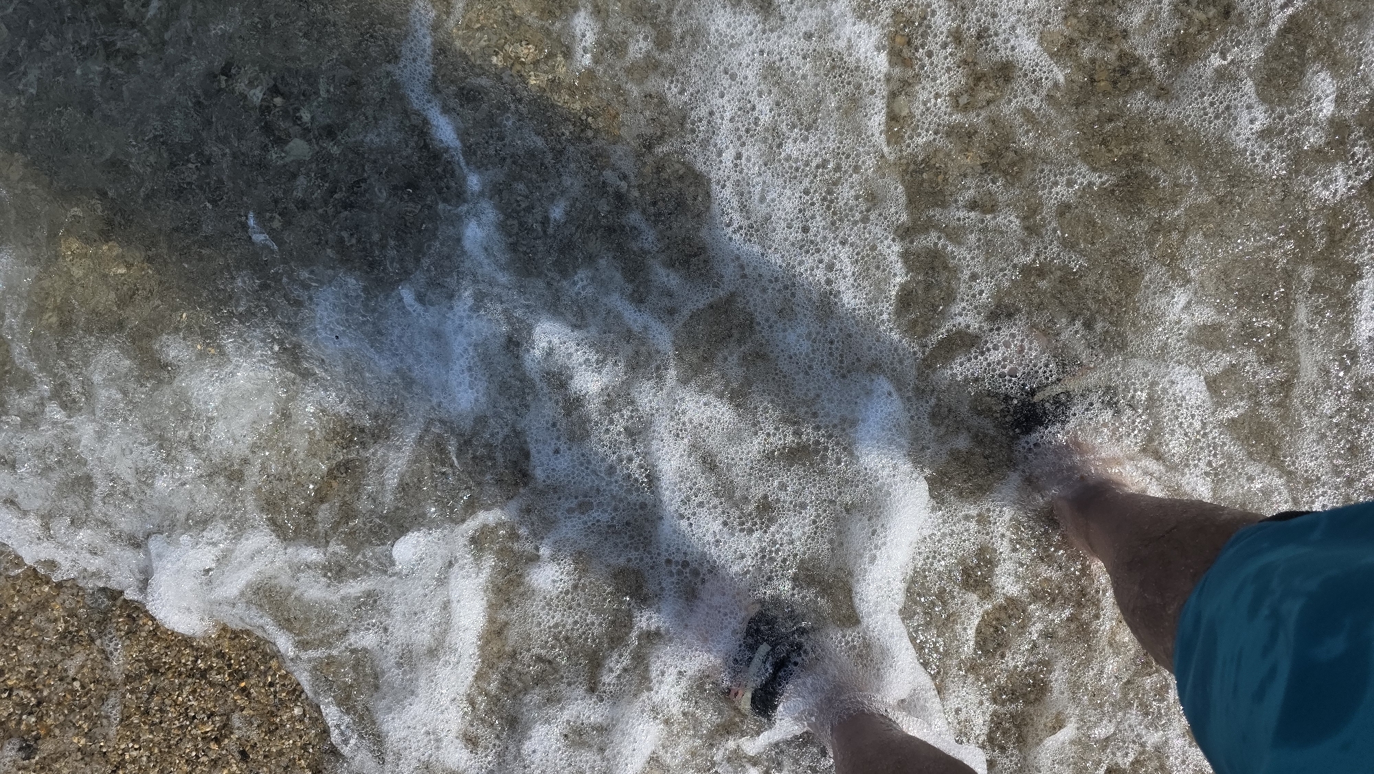 An overhead shot captures a person wading in the shallow, clear waters of a beach in Ασπροβάλτα, Greece. The lower torso and legs of an individual are visible, clad in blue swimming trunks. Dark-colored sandals or water shoes are worn on both feet, partially submerged in the foamy water. The water is highly agitated with extensive white foam and bubbles, indicating small waves breaking on the shore. Its transparency allows for clear visibility of the seabed. The visible shoreline on the left consists of light brown pebbles and gravel. Beneath the water, the seabed varies, showing a mix of lighter stones near the person's feet and darker, possibly deeper or rockier, patches further into the frame. Bright sunlight illuminates the scene, creating strong highlights and shadows on the water's surface.