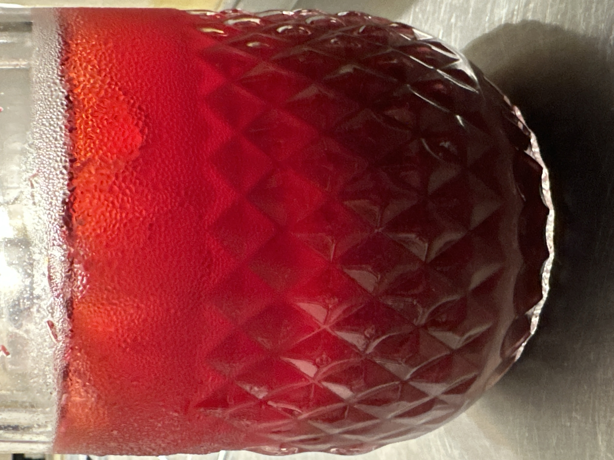 The image shows a close-up of a glass filled with a red liquid, likely juice or a cocktail. Condensation is visible on the outside of the glass. The glass has a textured, diamond-patterned surface. A shadow is cast on the surface the glass is resting on. There is no indication this image was taken in Новоглаголево, Russia.