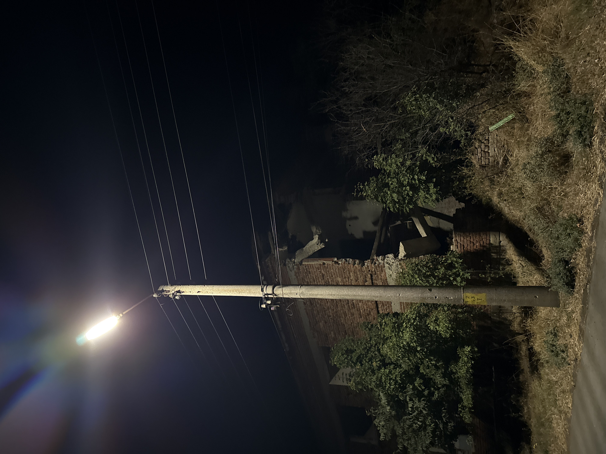The image depicts a night scene in the city of Ekzarh Antimovo, Bulgaria, featuring a street lamp illuminating a partially destroyed brick building and surrounding overgrown vegetation. The street lamp, positioned in the foreground, casts a bright light upwards and outwards, creating lens flares and illuminating a concrete utility pole with electrical wires stretching across the frame.
The building in the background shows significant structural damage, with large sections of its brick facade and interior missing or collapsed. The damage suggests a recent event, possibly an explosion or impact, that has left the building open to the elements. Green trees and bushes grow around the building, some of which appear to be encroaching on the structure itself. The ground is covered in dry grass, weeds, and some patchy greenery, indicating an unkempt or neglected area. A paved surface, likely a road or pathway, is visible in the lower left corner of the image. The overall atmosphere is dark and somber, with the artificial light of the street lamp providing the only significant illumination in the otherwise black night sky. There are no people or animals visible in the scene.