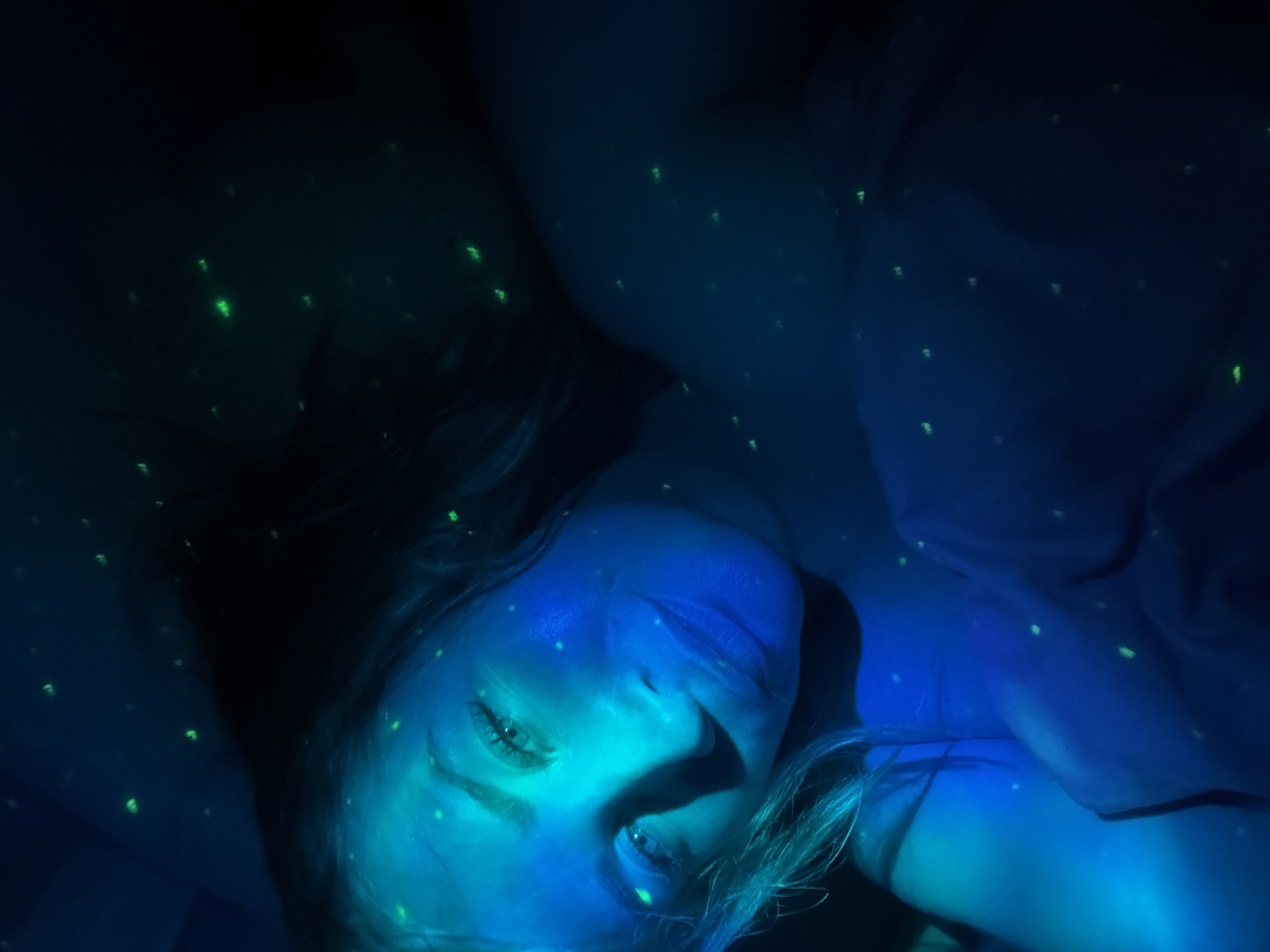 The image depicts a female individual reclining in a dimly lit indoor setting, predominantly illuminated by a blue light source. Her face and upper torso are visible, with her head turned slightly towards the viewer. She possesses light-colored hair and is looking directly into the camera. Her skin and a portion of her garment, which appears to be light-colored fabric, are covered in numerous small, distinct, luminous green speckles or dots. These green elements are also distributed across the dark background, suggesting a widespread application or projection. The environment is characterized by deep shadows and a general lack of ambient light, consistent with an interior space. This scene is situated in the city of Veliky Novgorod, Russia.