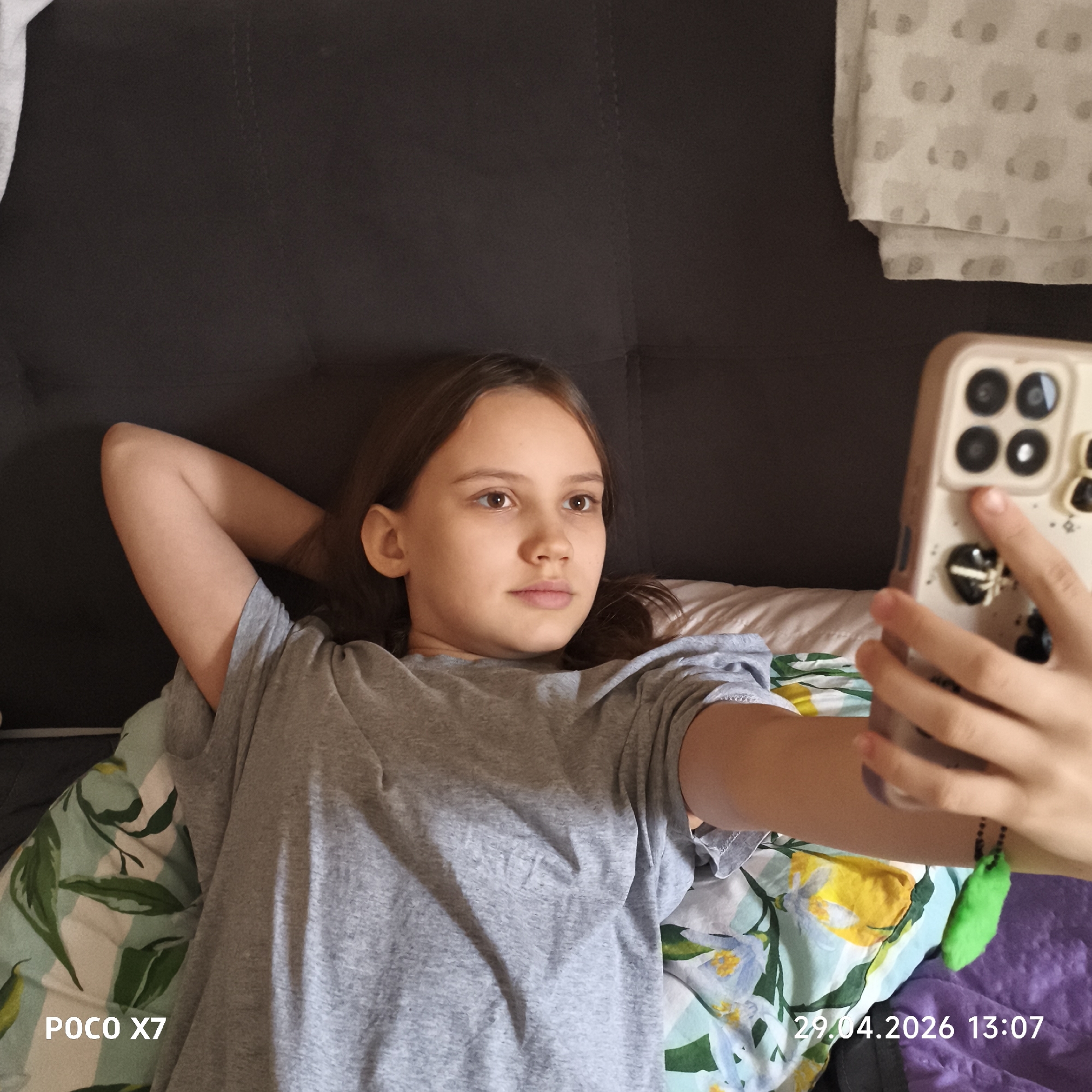 The image captures a young female indoors, likely in Chaikovsky, Russia, on April 29, 2026, at 13:07. The subject is lying down on a bed or sofa, wearing a plain grey t-shirt. Her brown hair is down, and her arms are relaxed behind her head. She is holding a light-colored smartphone with her right hand, oriented towards herself, suggesting she is taking a selfie or engaging with the device. Her gaze is directed towards the phone's screen, and her expression is neutral and calm.
The smartphone case features a distinct design with multiple camera lenses arranged in a square pattern and decorative elements including small black heart shapes and a vibrant green, fluffy charm attached to the wrist strap.
The setting appears to be a comfortable indoor space, possibly a bedroom or living room. Various soft furnishings are visible: a dark grey or brown upholstered sofa or headboard behind her, a patterned pillow or duvet with green leaves and yellow and blue flowers beneath her, a white patterned sheet with light grey bear-like figures in the upper right, and a purple blanket or fabric in the bottom right.
Visible text in the image includes "POCO X7" in the bottom left corner, indicating the device used to capture the photo, and the date and time "29.04.2026 13:07" in the bottom right corner. The lighting is soft and ambient, typical of an indoor daytime setting.