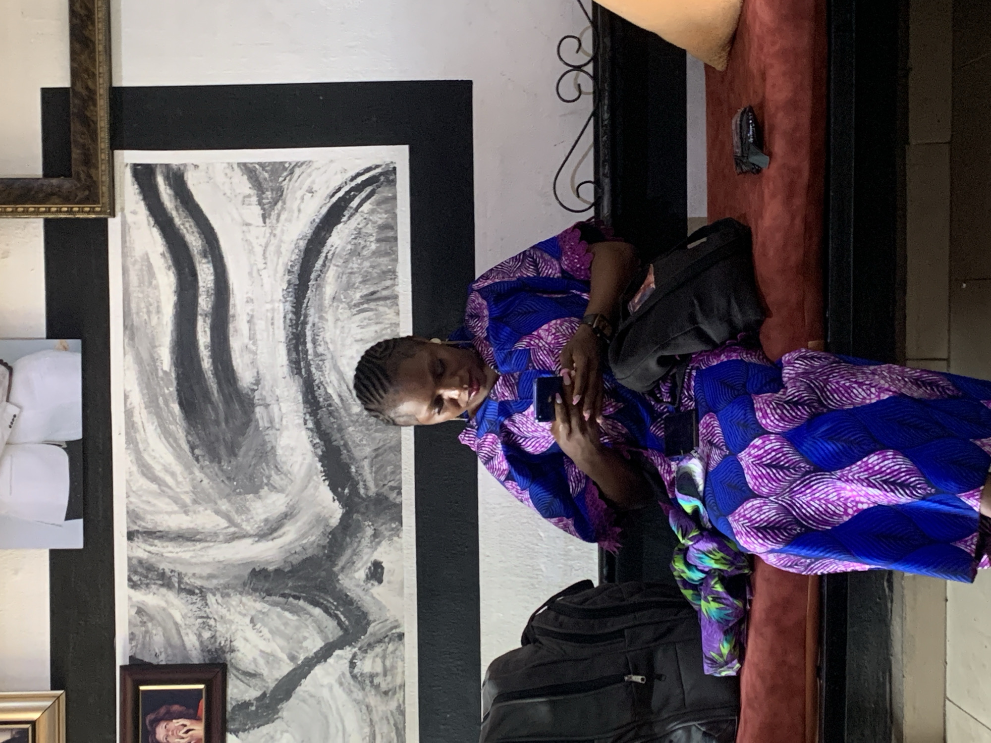 A woman sits on a dark red couch in what appears to be an indoor waiting area or lounge. She wears a vibrant blue and purple patterned dress and is looking at a cell phone in her hands. Beside her is a black backpack, and a black purse rests on the couch near her. The room's wall is adorned with a large black and white abstract painting, and several framed photos are visible. The setting is likely in Gura Top, Nigeria, based on the prompt.