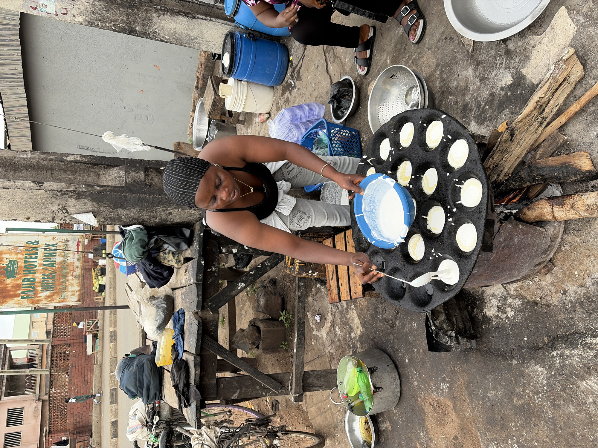 A woman in a black tank top and gray sweatpants pours batter onto a hot, round griddle with circular indentations in an outdoor setting, presumably in Jos, Nigeria. The griddle rests over a fire fueled by wood, suggesting a street food vendor setup. She uses a spoon to transfer batter from a blue and white bowl to the hot surface. Several containers and utensils are scattered around her, including blue barrels, buckets, and metal bowls. A second person is visible in the background, seated on one of the blue barrels. A bicycle is parked nearby, partially obscured by bags and containers. A faded sign reading "FAJIB HOTELS & NITES/ANNEX" is visible on a building across the street, further suggesting an urban, possibly commercial, location. The ground is concrete and appears worn and stained.