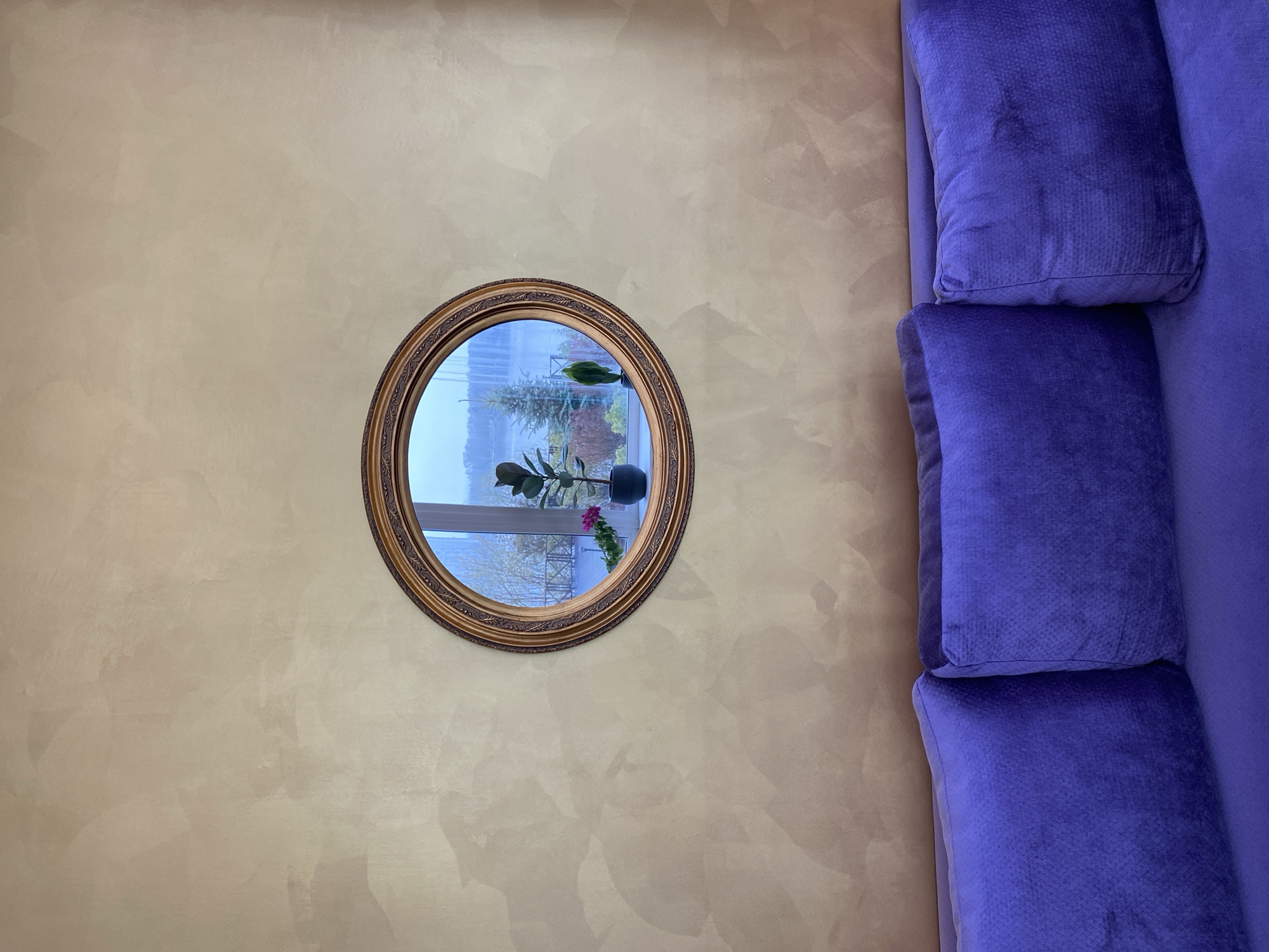 The image shows a portion of a wall with a round mirror and part of a purple sofa. The wall is decorated with a mottled, textured finish in shades of beige and tan. The mirror has an ornate, gold-colored frame. The reflection in the mirror reveals a window with plants on the windowsill and a view of the outside, with trees and what appears to be a metal structure. To the right of the mirror is a blue sofa with a textured surface, and on top of it, there are three cushions. There is no confirmed evidence to suggest that the image was taken in Yekaterinburg, Russia.