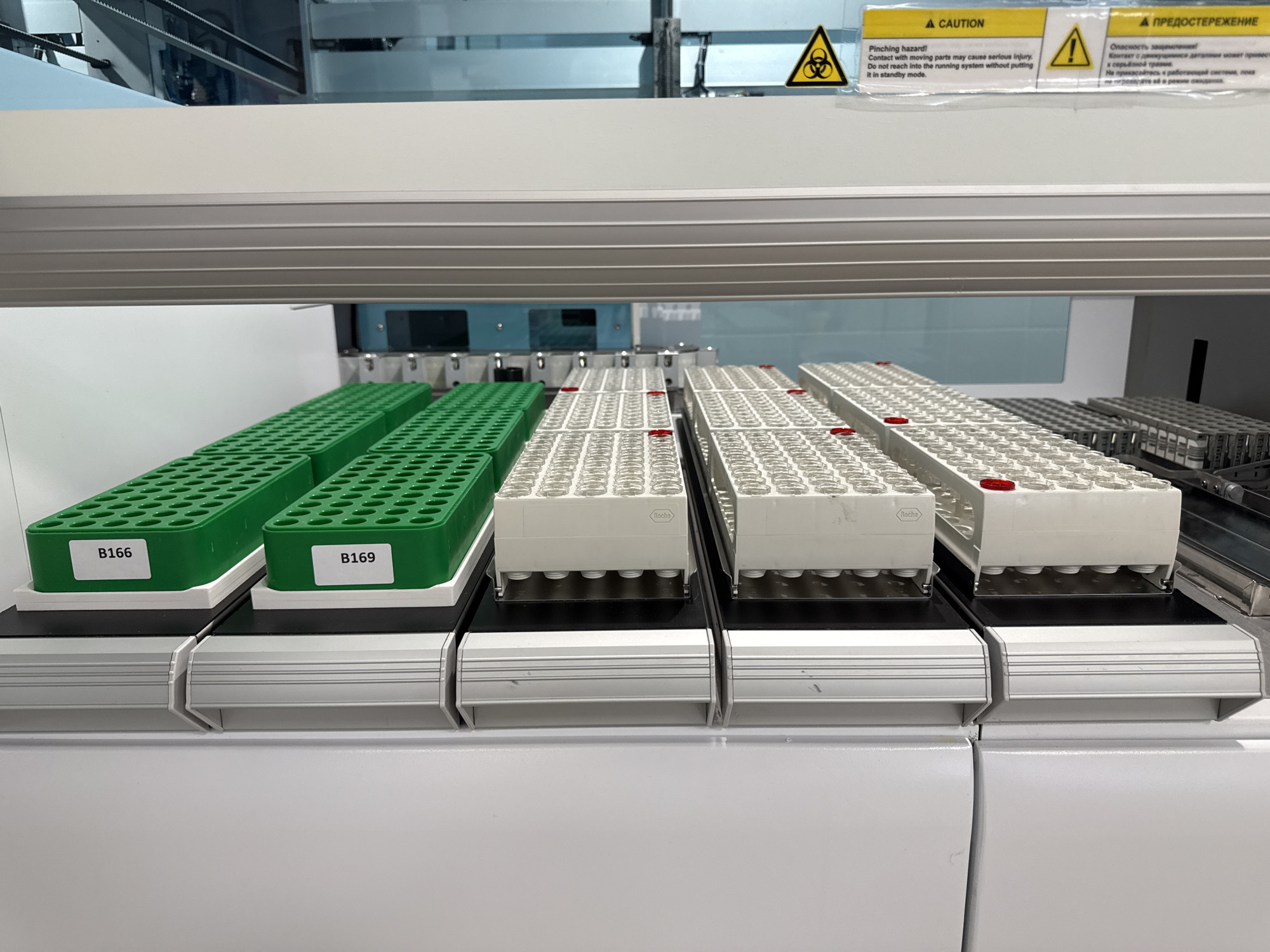 The image shows a laboratory setting, featuring an automated analyzer. Two green racks labeled "B166" and "B169" stand to the left, filled with test tubes. Three white racks filled with test tubes and labeled with the brand name "Roche" are positioned next to them. A warning sign is placed above the racks, stating "CAUTION Pinching Hazard!" in both English and Russian. The environment appears to be clean, well-lit, and organized. There is no evidence to suggest this scene is in Moscow, Russia.