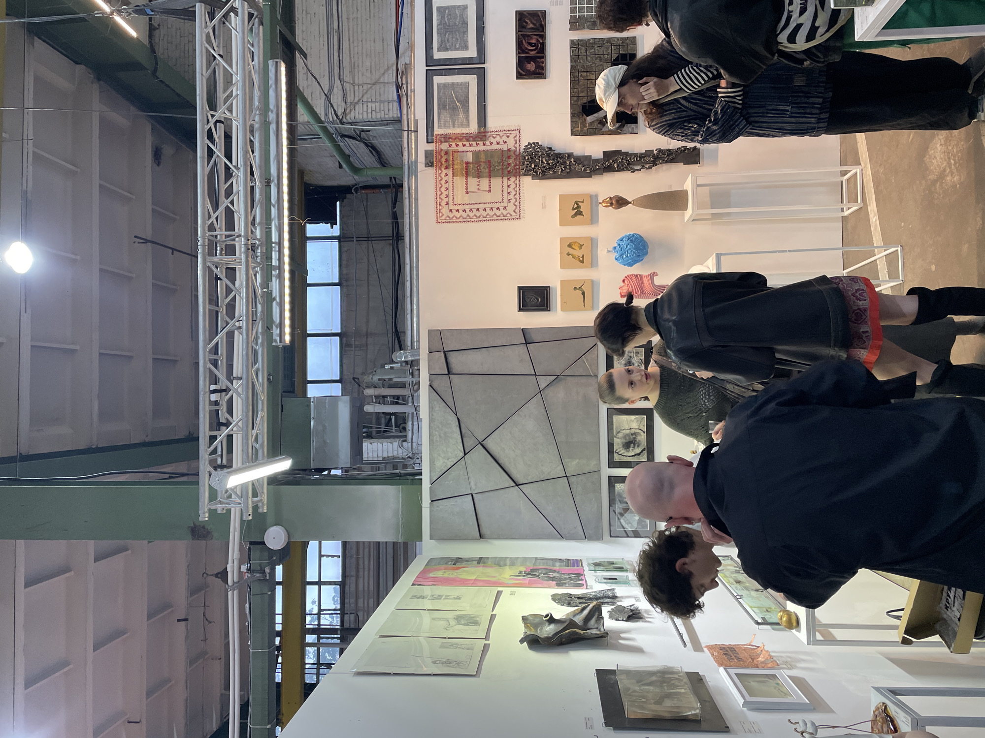 The image shows an indoor art exhibition with multiple artworks displayed on white walls. Several people are viewing the art. The space has high ceilings with visible metal support structures and lighting fixtures. Windows are visible in the background. The location may be an industrial space or warehouse converted into an exhibition venue, but city information is not available from the image.