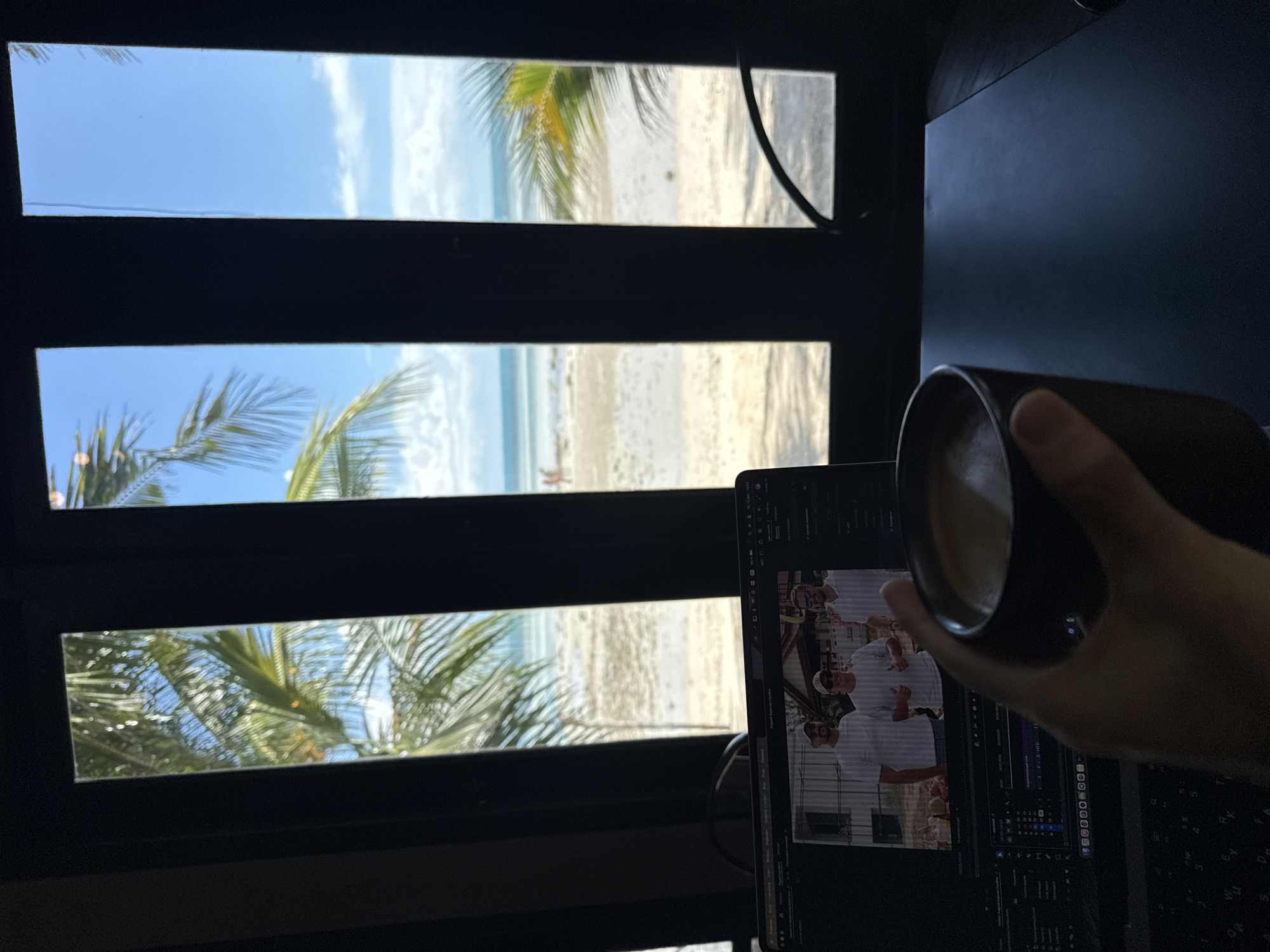 A person's hand holds a dark mug of coffee in the foreground, obscuring part of a laptop screen. The laptop displays a video editing timeline and a still image of three men wearing white shirts and sunglasses. Behind the laptop, a window is divided into vertical panes by dark framing. Through the window, a tropical beach scene is visible, with palm trees, sand, the ocean, and a clear blue sky with scattered clouds. The location is Ko Samui District, Thailand, suggested by the tropical vegetation and beach. The overall impression is of someone working or relaxing in a scenic, tropical environment.