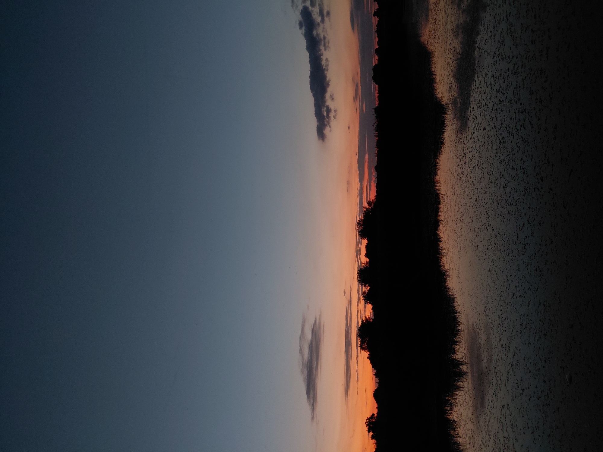 A silhouette of trees and vegetation lines the horizon, reflected in a dark body of water. The sky above is a gradient of deep blue to a soft orange hue near the horizon, indicating either dawn or dusk. A few wispy clouds are scattered across the sky. The scene appears to be in a natural, rural setting, possibly near Ustyvytsia, Ukraine, given the context. There are no visible people or animals.