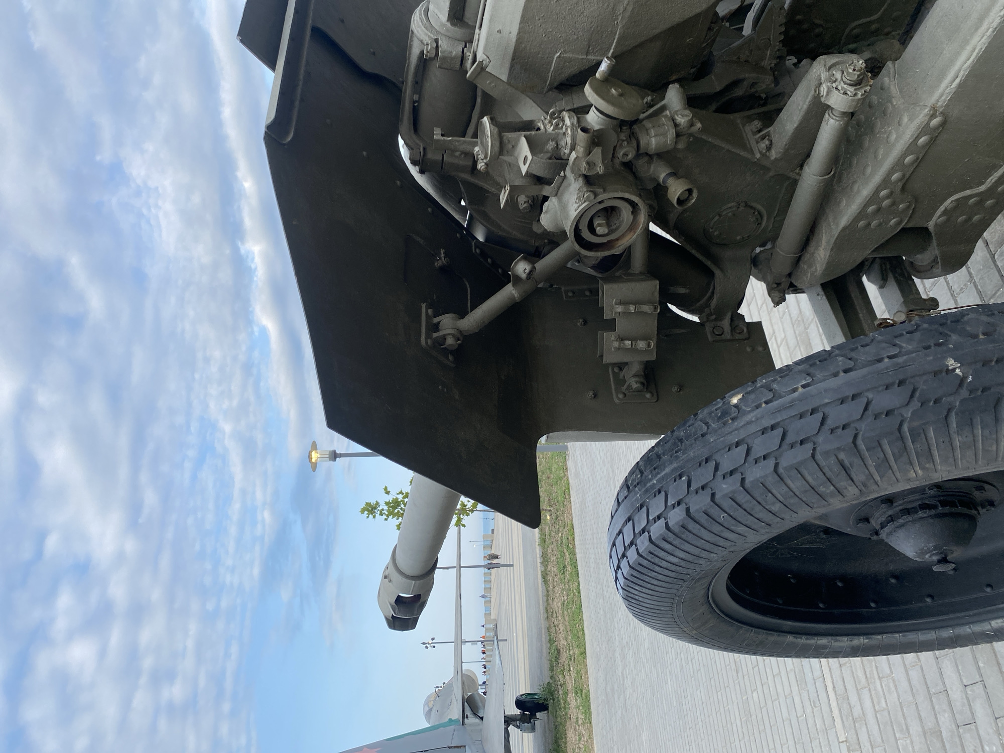 The image shows a close-up of a military cannon's wheel and part of its mechanism, with an airplane in the background. The cannon appears to be olive-drab green, as is the airplane. The scene is outdoors on a paved area, likely a promenade, with a body of water visible in the distance. The sky is partly cloudy, with a lamppost visible in the background. Several people are walking in the distance, appearing as small figures on the promenade. The location is identified as Arablyar, Russia.