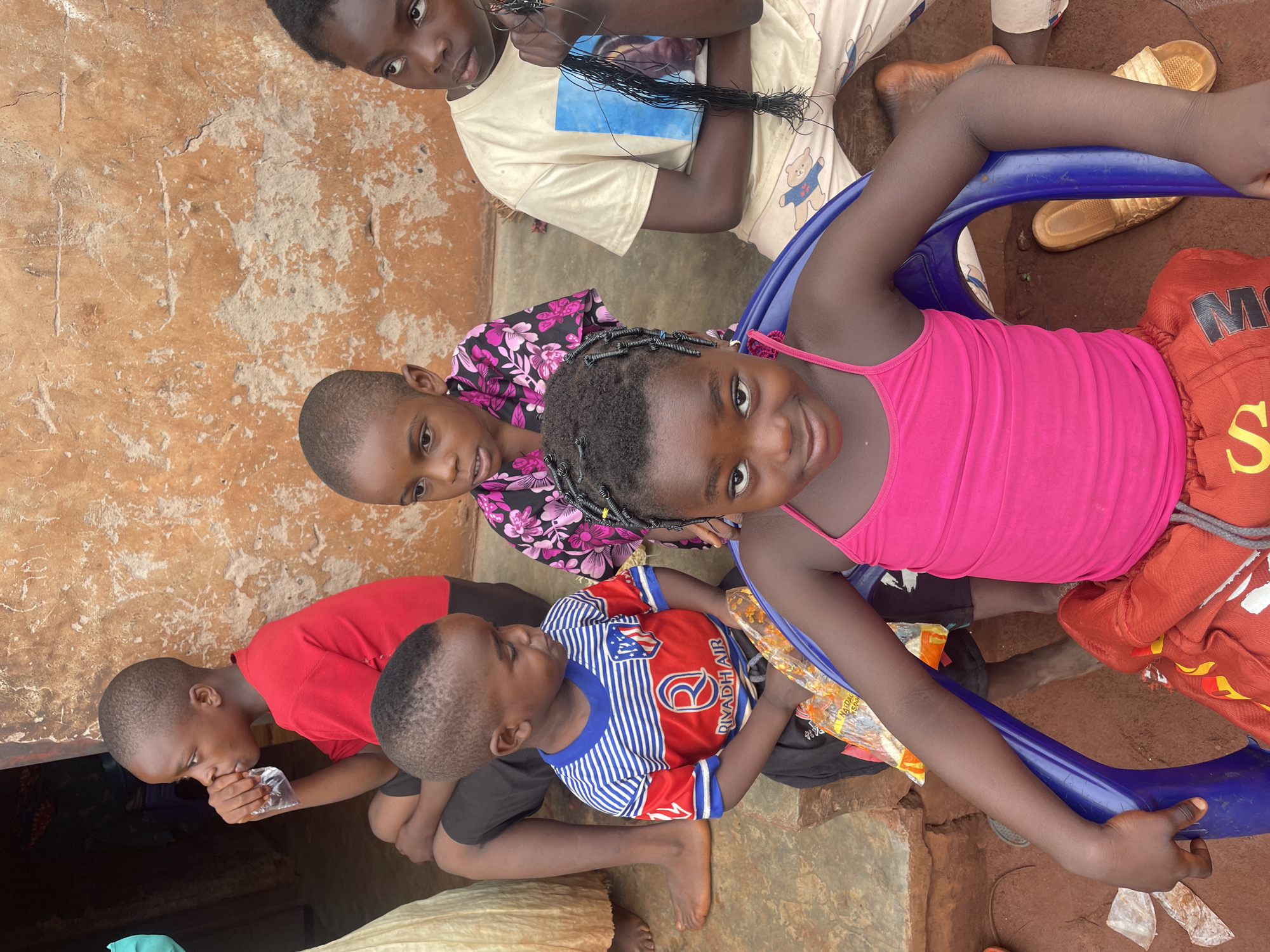 A group of six children are gathered outdoors in what appears to be the city of Ogbe, Nigeria, likely in a residential area. The background features a wall or structure with an earthy, possibly mud-based surface. The foreground is a dirt ground. The children are positioned in a cluster, some seated and others crouching. One child is wearing a pink tank top and sitting on a blue plastic chair. Several of the children are looking towards the camera. A boy in a red and blue striped shirt is seated with another child on his lap. A boy in a red shirt appears to be drinking from a plastic bag. Another child is wearing a shirt with a floral print. A child near the top of the image is seated with a collection of hair extensions in their hands. Several pairs of sandals are visible in the vicinity of the children. Plastic wrappers and debris are scattered around the ground.