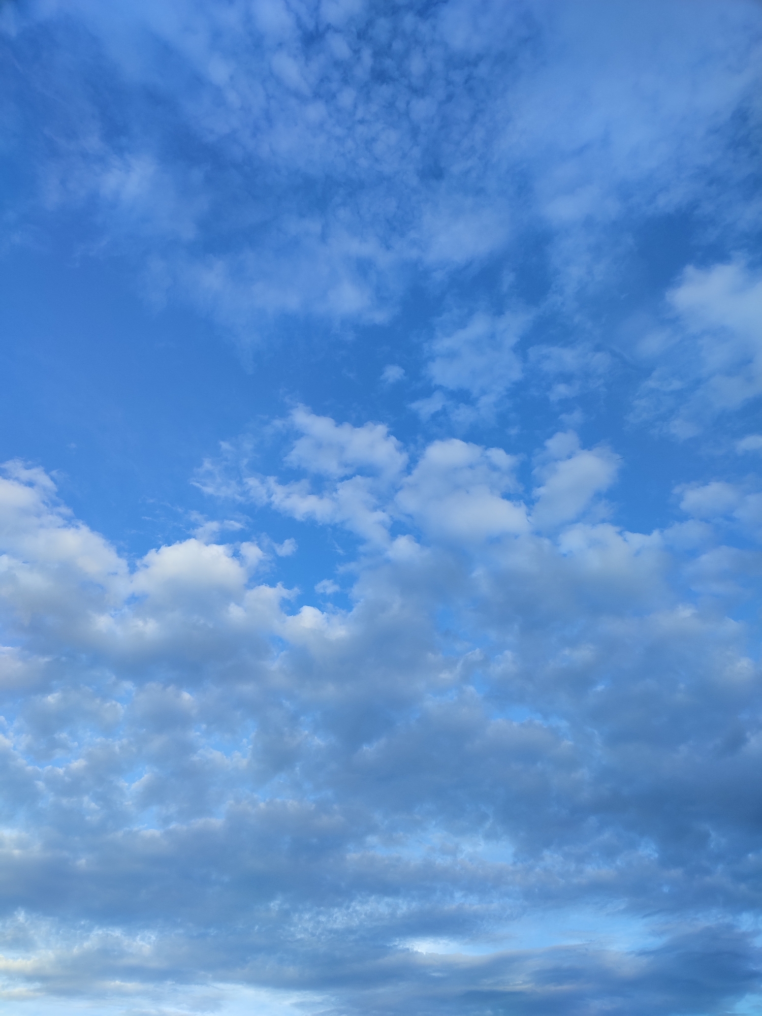 The image shows a clear blue sky with scattered clouds over the city of Voskobiinyky, Ukraine. The clouds are a mix of wispy cirrus clouds higher up and more defined cumulus clouds lower down, some of which appear to be casting shadows. The lighting suggests it is daytime, possibly late afternoon given the slightly diffused light and the variations in cloud brightness. There are no visible people, animals, or man-made structures in the frame. The focus is entirely on the atmospheric conditions.