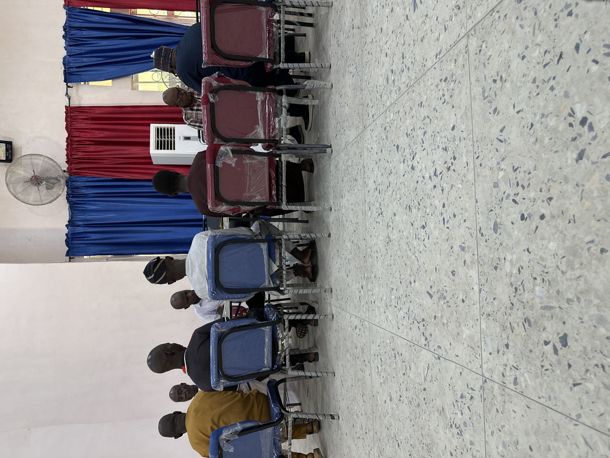 This media file shows a group of men sitting in chairs in what appears to be a meeting or gathering. The scene is indoors, with a light-colored wall and a tiled floor. The chairs are a mix of red and blue, with some covered in plastic. Behind the chairs, red and blue curtains are visible. An air conditioner unit is mounted on the wall, and a fan is also present. The lighting suggests it could be daytime. The men are dressed in various forms of attire, including traditional clothing and shirts. Some are wearing head coverings. The overall atmosphere seems calm and focused. The location is identified as Jalingo, Nigeria, but no specific landmarks or textual information are visible to further pinpoint the exact location within the city.