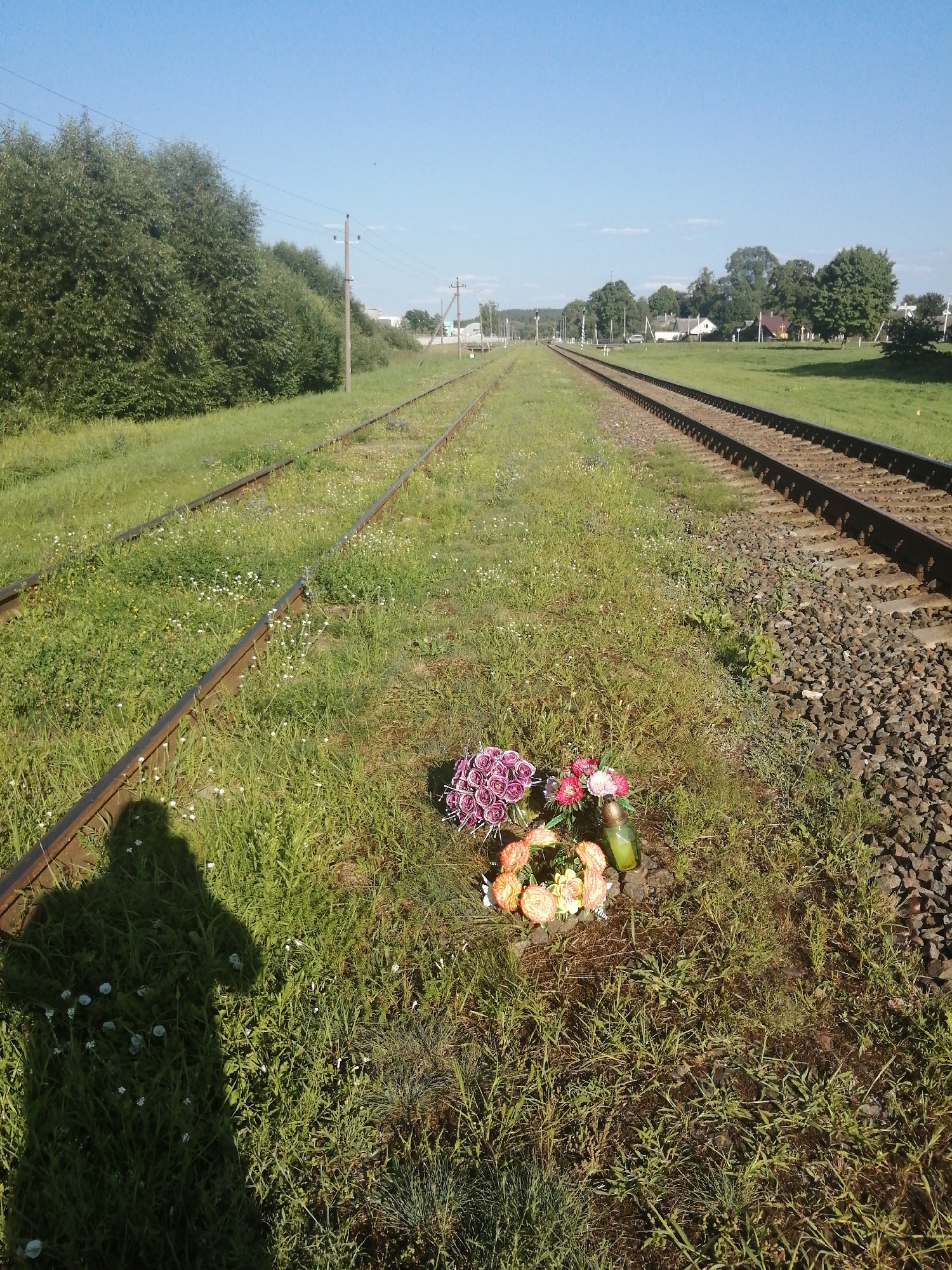 The photograph captures a view along a rural railway line. The tracks themselves, made of weathered steel, are flanked by overgrown green grass and small white wildflowers. In the foreground, on the left side of the tracks, a dark shadow of a person is cast onto the grass. Slightly to the right of the shadow, and closer to the center of the frame, a floral wreath and a green glass candle holder are placed on the ground. The wreath is composed of artificial flowers in shades of purple, pink, and orange, with some smaller yellow flowers. In the background, the railway tracks recede into the distance, with power poles and overhead wires visible along the right side of the tracks. Beyond the tracks, there are trees and a few houses, suggesting a small settlement or village. The sky is clear and blue. This scene is located on the outskirts of the city of Slonim, Belarus, near the former railway station of Al'khovtsy.