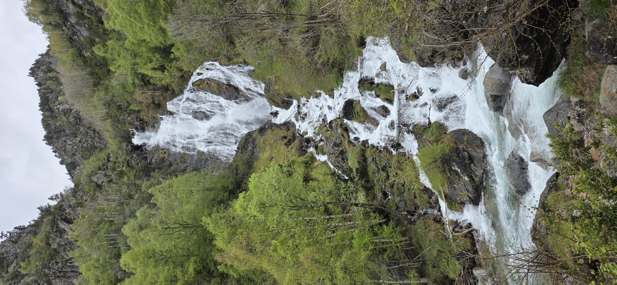 The image shows a cascading waterfall in a mountainous region. The location is likely near Sahún, Spain, based on the prompt.
The waterfall is the primary subject, with water flowing rapidly over rocks and down the mountainside. The surrounding environment includes green vegetation covering the slopes, with a mix of trees and grassy areas. The sky appears overcast.
There are no people or other subjects readily visible in the scene. The focus is entirely on the natural landscape.