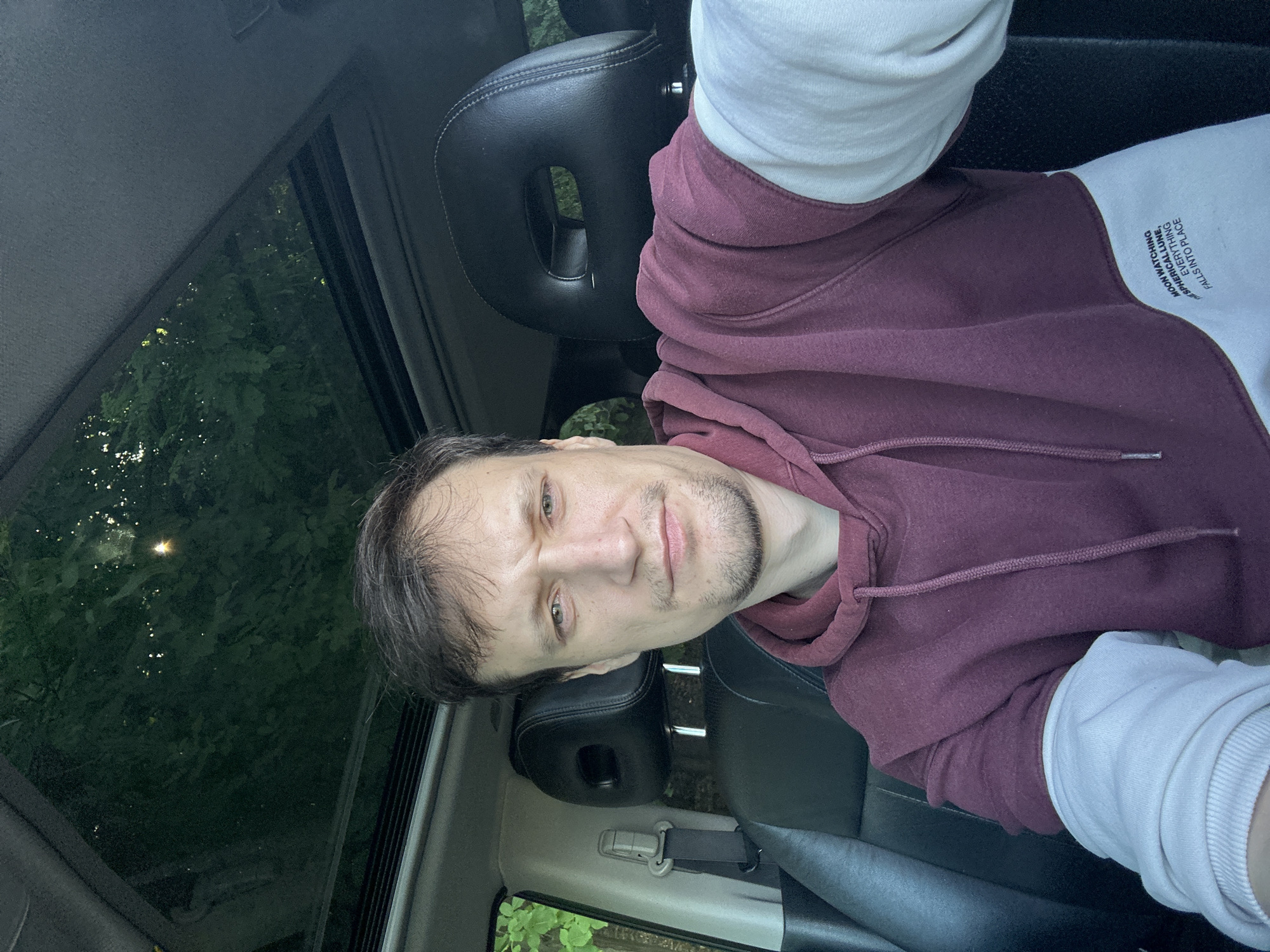 A man with short, dark hair and a goatee is sitting in the driver's seat of a car, looking at the camera. He is wearing a burgundy hoodie over a white long-sleeved shirt. The car has black leather seats and a sunroof, through which trees and a bright sky are visible. The man's left hand is visible, resting on his lap. The image is a selfie, taken from the perspective of the person behind the camera. The location is not specified in the image.