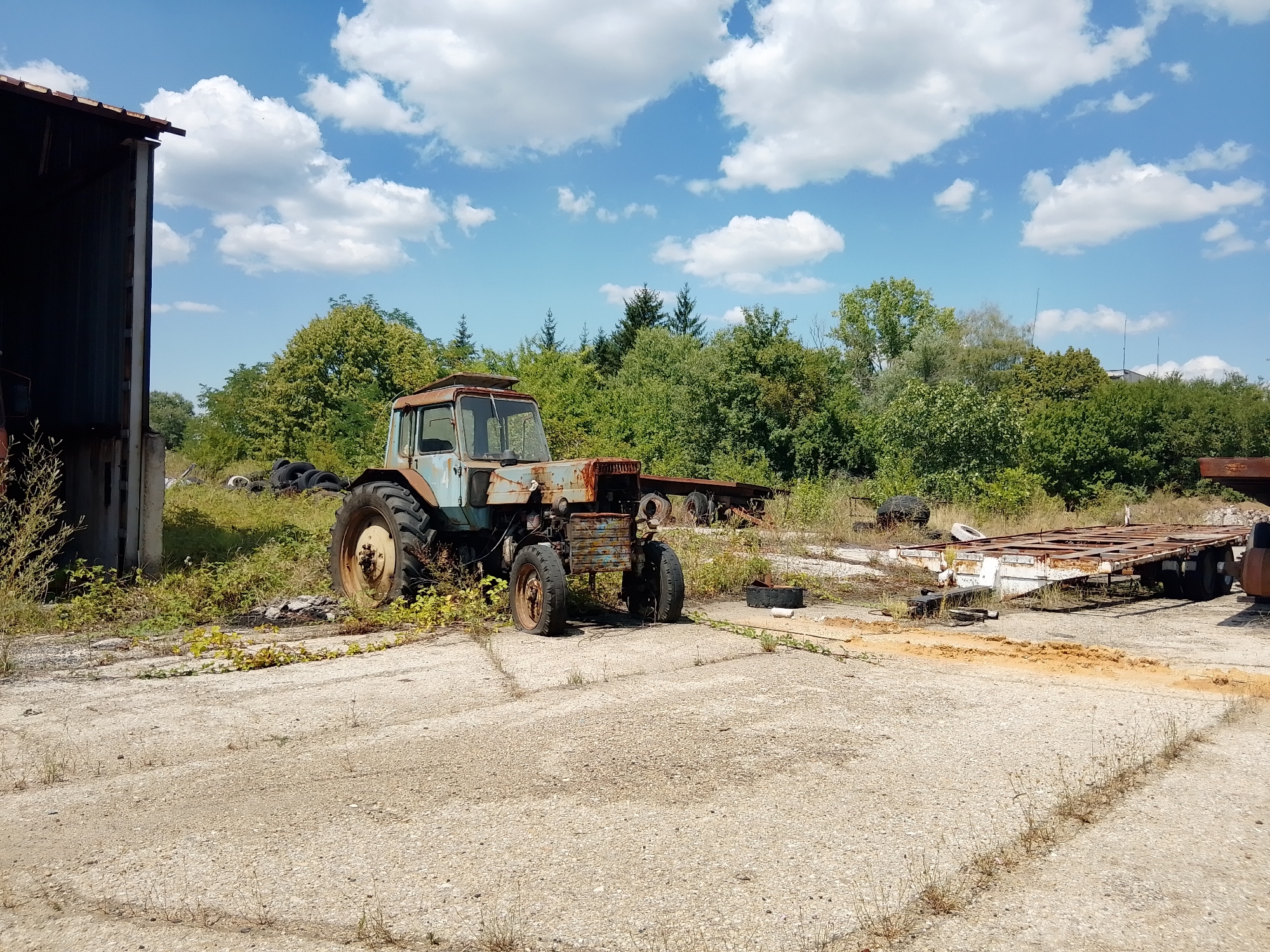 An abandoned tractor and several flatbed trailers are parked on a gravel lot next to a dilapidated building under a partly cloudy blue sky. The tractor is light blue and rusted, with large, weathered tires. It is positioned in the foreground, slightly to the left of center. Behind the tractor, a long, rusty flatbed trailer with multiple axles is visible, along with another smaller trailer. Tires are scattered on the ground near the tractor. The building on the left appears to be made of corrugated metal. Trees and brush form a dense backdrop behind the vehicles. The ground is a mixture of gravel and cracked concrete, with sparse vegetation growing in patches. The scene suggests a junkyard or an area for storing old machinery.