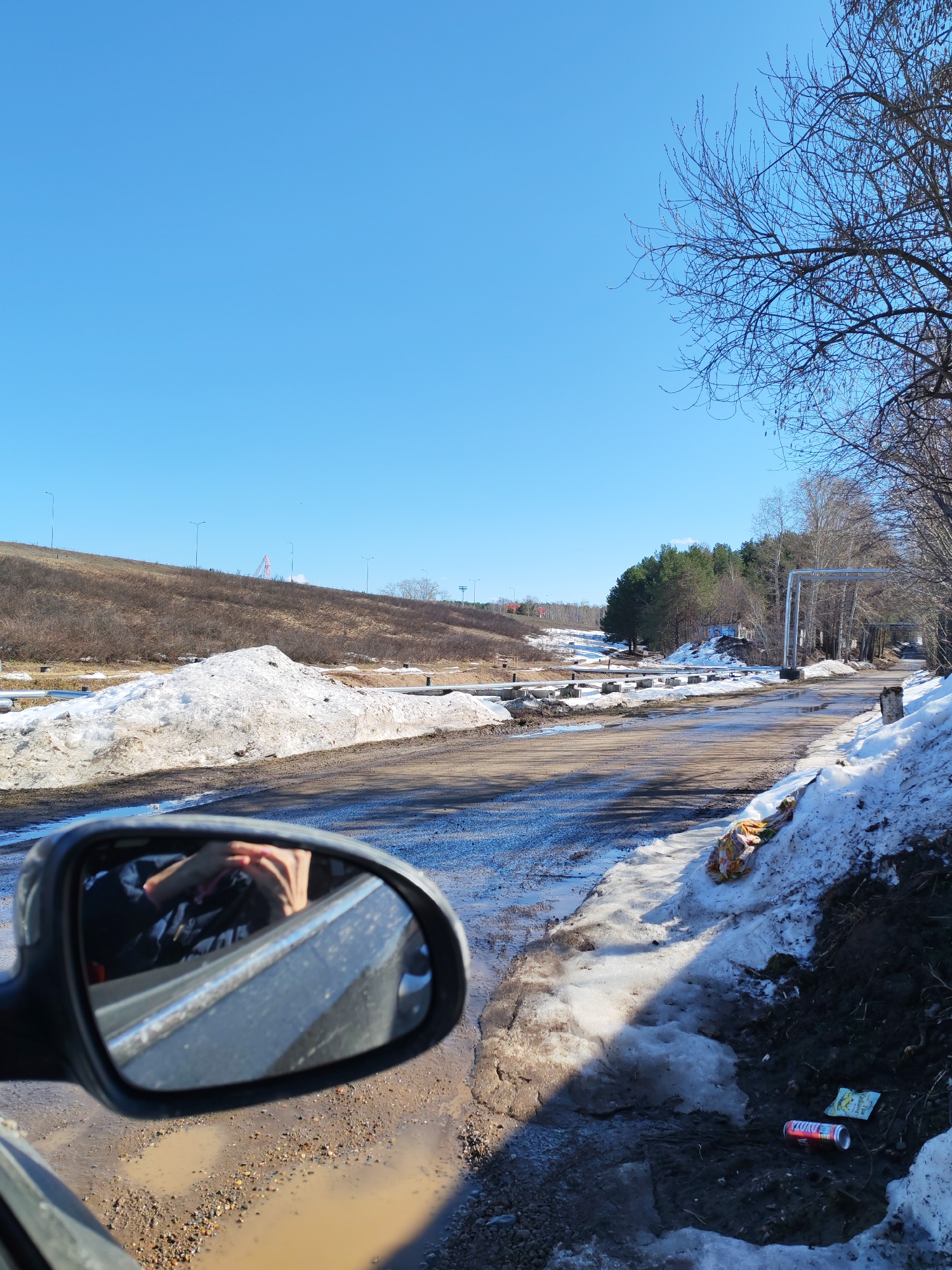 The image depicts a muddy road in Chaikovsky, Russia during a thaw with remnants of snow on either side. The clear, blue sky suggests a sunny day. Bare trees line the right side of the road. In the distance, a grassy hill is visible with lampposts and what seems to be a pink structure on top. A metal structure spans the road further ahead. The reflection in the side mirror of a car shows an arm and a jacket in the car. Litter, including a can and wrapper, are visible along the roadside on the right.