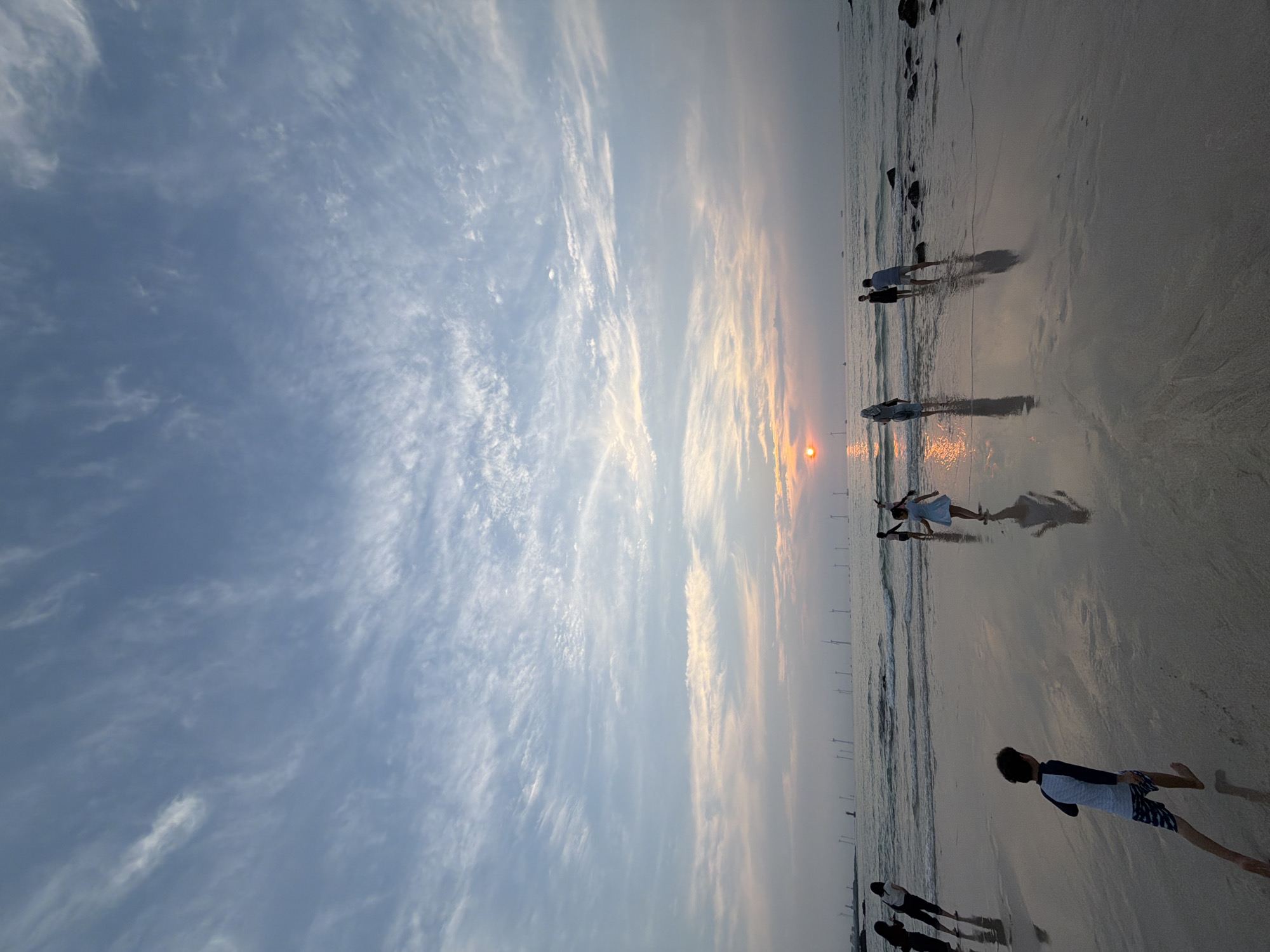 The image depicts a serene beach scene on Jeju Island, South Korea, at sunset. The sky is adorned with wispy clouds, transitioning from a soft blue to hues of orange and pink as the sun dips towards the horizon. In the distance, a line of wind turbines stands tall against the water, their silhouettes stark against the fading light.
The sandy beach is wet and reflective, mirroring the sky and the figures present. Several people are scattered across the shoreline and in the shallow water. In the foreground, a young boy in a swimsuit runs towards the viewer, his reflection stretched out on the wet sand. Further back, a girl in a light-colored dress walks near the water's edge, her reflection also visible. Other individuals are seen wading in the sea or standing on the beach, some appearing to interact with each other. The water itself is calm with gentle waves lapping the shore. The overall atmosphere is one of tranquility and natural beauty, characteristic of Jeju's coastal landscapes.