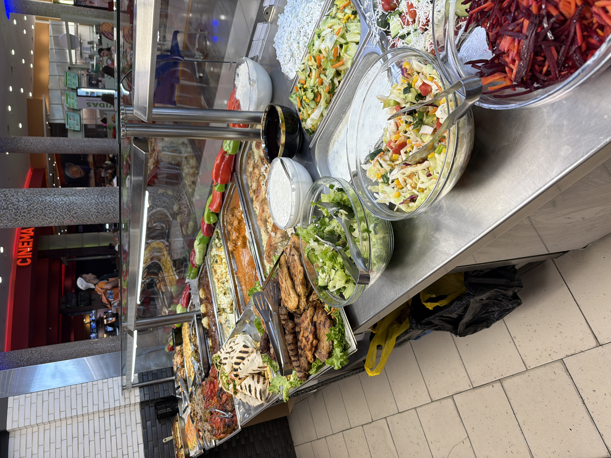 The image depicts a food buffet with a variety of dishes, likely in a casual dining setting. The buffet is organized in stainless steel trays and glass bowls, showcasing an assortment of cooked meats, grilled items, rice, pasta, and several types of salads. Visible are grilled chicken, seasoned meats, flatbreads, lentil dishes, a white rice dish, and various mixed vegetable salads including what appears to be a coleslaw and a grated beetroot and carrot salad. There are also bowls of what looks like yogurt or sour cream dips.
The buffet is situated within a larger establishment, indicated by the presence of a "CINEMA" sign in red lettering in the background, suggesting a location within a mall or entertainment complex. The overall environment appears to be a food court or a cafeteria area. In the background, out of focus, are more seating areas and possibly other food vendors. The flooring is tiled with a beige-toned tile. On the left side of the frame, a brick wall with white grout is visible. Underneath the buffet counter, there are black and yellow trash bags.
While the specific location in Plovdiv, Bulgaria is not discernible from the visual information alone, the presence of the "CINEMA" sign suggests a modern commercial area. The style of the buffet and the general ambiance are consistent with many food courts found in shopping malls in urban centers.
