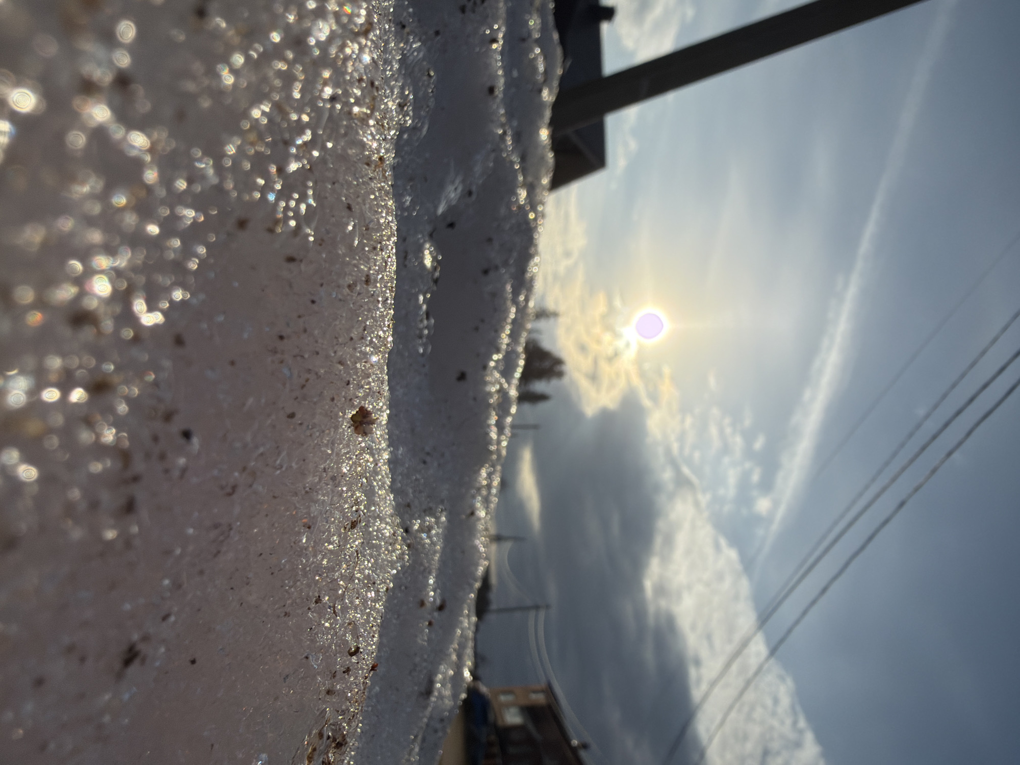 The image, taken in Chaikovsky, Russia, captures a close-up, low-angle perspective of a sparkling snow or ice bank against a bright sky. The original image appears rotated 90 degrees clockwise; when viewed upright, the snow/ice forms a prominent bank along the left side of the frame, stretching towards the viewer. This snow or ice is highly textured and glitters intensely due to direct sunlight, exhibiting a bokeh effect with circular light reflections, some showing rainbow diffraction. Small pieces of debris, possibly dirt or grit, are embedded within the icy surface.
In the background, the sky dominates the right side of the image. The sun, a bright, slightly purple-tinged orb, is low on the horizon, creating a strong glare and suggesting either sunrise or sunset. Various clouds, ranging from wispy to more substantial, are scattered across the sky, partially obscuring the sun and creating dramatic light patterns. Overhead, three parallel power lines extend diagonally across the upper right portion of the sky. A dark, indistinct structural element, possibly part of a building or utility pole, is visible near the top of the frame. Further in the distance, below the horizon, blurred shapes suggest buildings or structures, indicating a semi-urban or suburban setting.
No people are visible, and no specific activity is taking place beyond the natural interplay of light and ice. The scene conveys a serene winter moment, characterized by the bright, cold sparkle of the ice contrasting with the warm glow of the sun. There is no visible text.