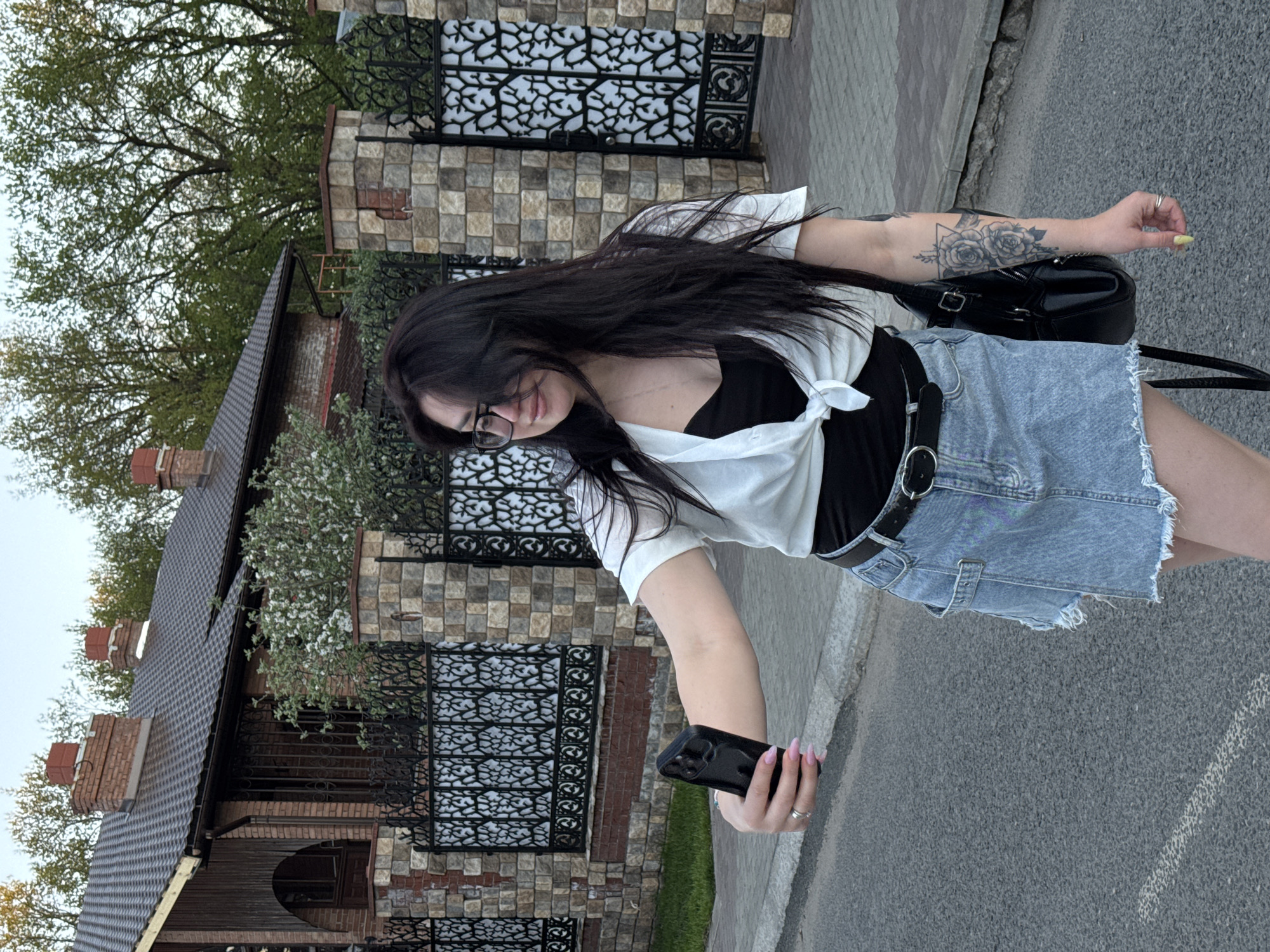 A woman with long, dark hair, wearing glasses, a black top, a white shirt tied at the waist, and a denim skirt is holding a black phone in her left hand, presumably taking a picture or video. She has a tattoo on her left arm. A black backpack is slung over her shoulder. She is standing on a paved road. Behind her is a stone and metal fence, and a house with a brown roof and brick chimney. The setting appears to be an urban or suburban environment. The location is Unknown, Unknown.