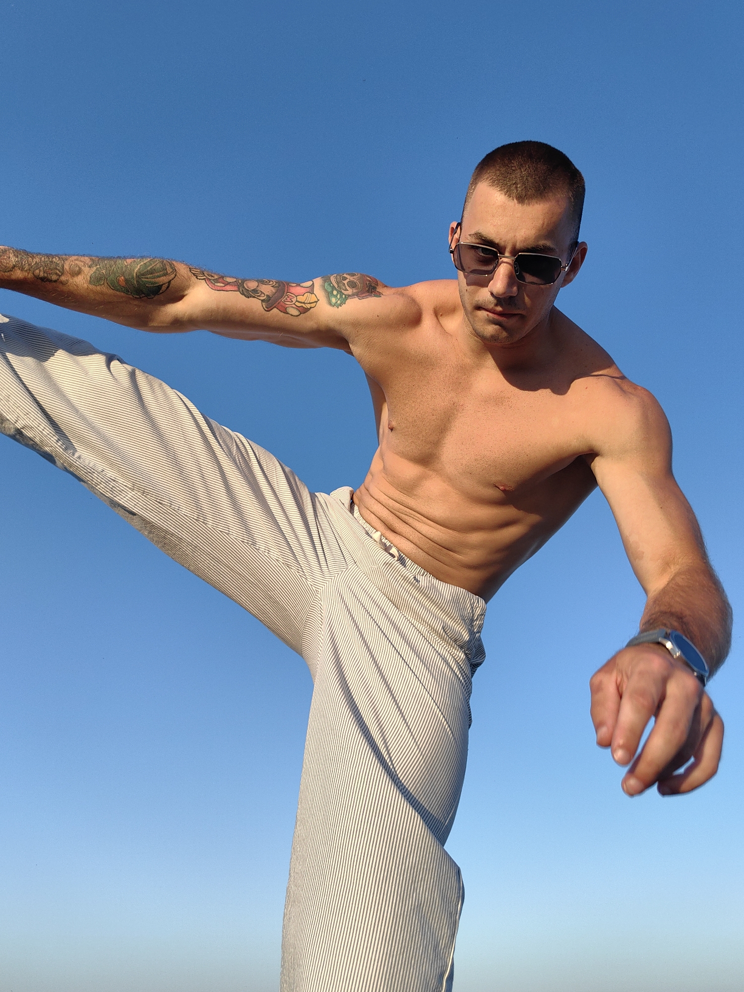 A shirtless man with short hair and sunglasses is captured in a dynamic pose against a clear blue sky. He is wearing striped pants and has tattoos on his left arm. The man appears to be mid-movement, with his left leg extended upwards and his right arm reaching down. A watch is visible on his right wrist. The lighting suggests it is daytime, and the cloudless sky indicates fair weather. The scene is likely outdoors, possibly in Varna, Bulgaria, given the context. The man's expression is focused, and his posture conveys a sense of athleticism or performance. There is no visible text in the image.