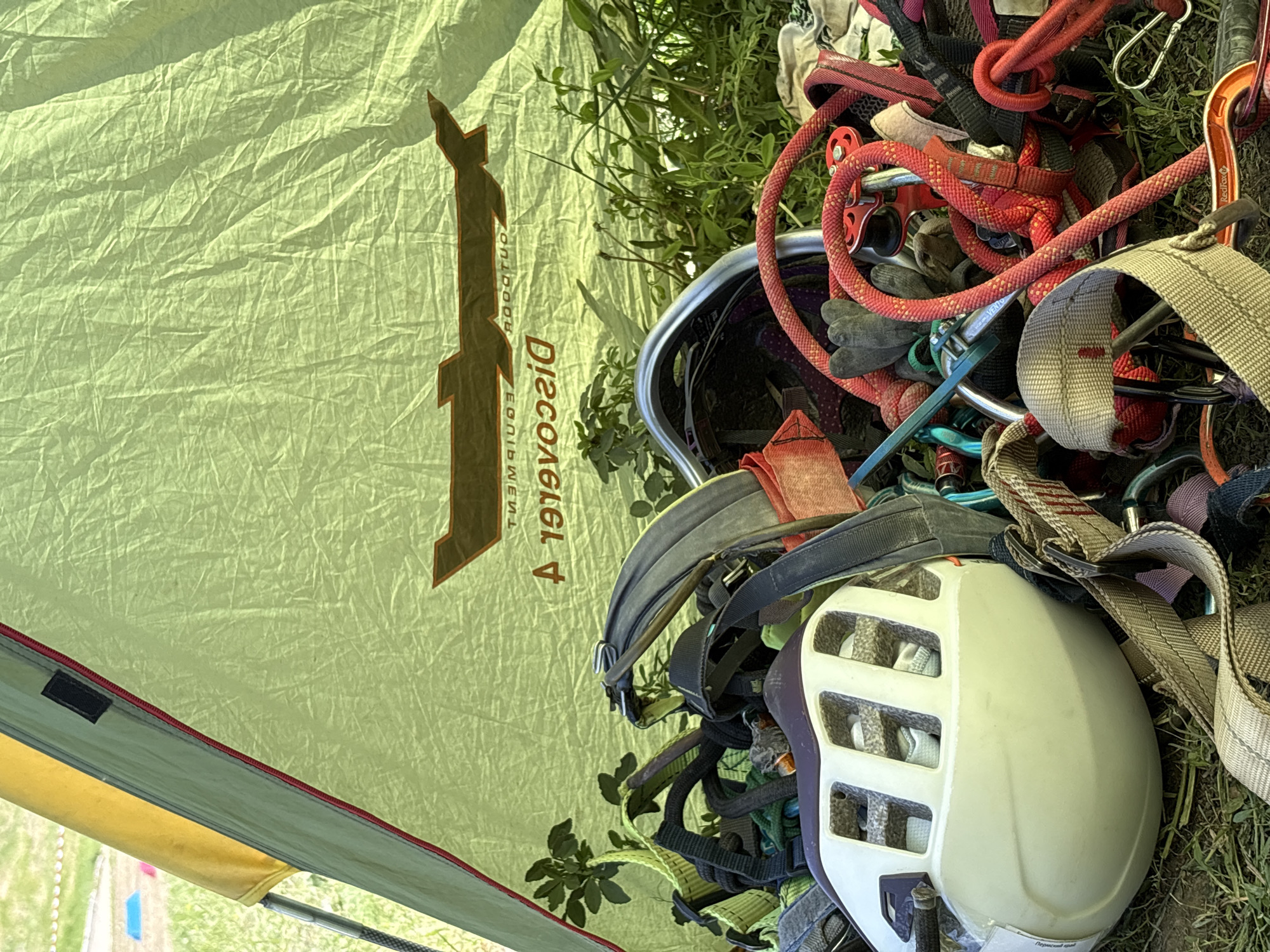 The image shows a tent and climbing gear. The tent is green with the logo "Discovery Equipment" printed on the side. Climbing gear, including helmets, ropes, and carabiners, is spread around the base of the tent. The location is not discernible from the image.