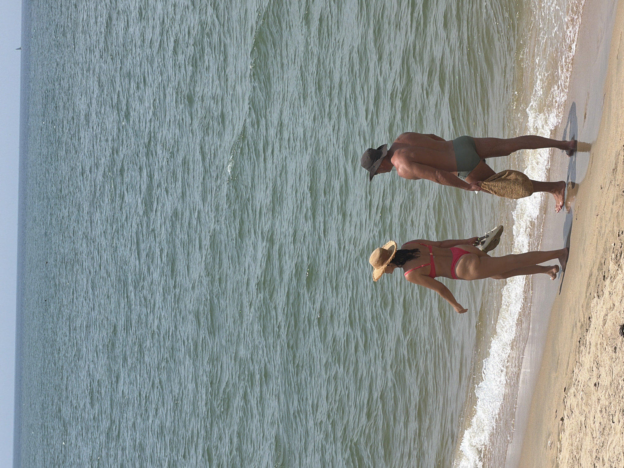 The image shows a woman and a man walking on a sandy beach in Nha Trang Ward, Vietnam, with the ocean in the background. The woman is wearing a pink bikini and a straw hat, and she is carrying a pair of white sandals in her right hand. The man is wearing dark swim trunks and a dark hat, and he is carrying a woven bag in his left hand. The ocean is blue-green with small waves. The sky is pale blue. The sand is light brown.