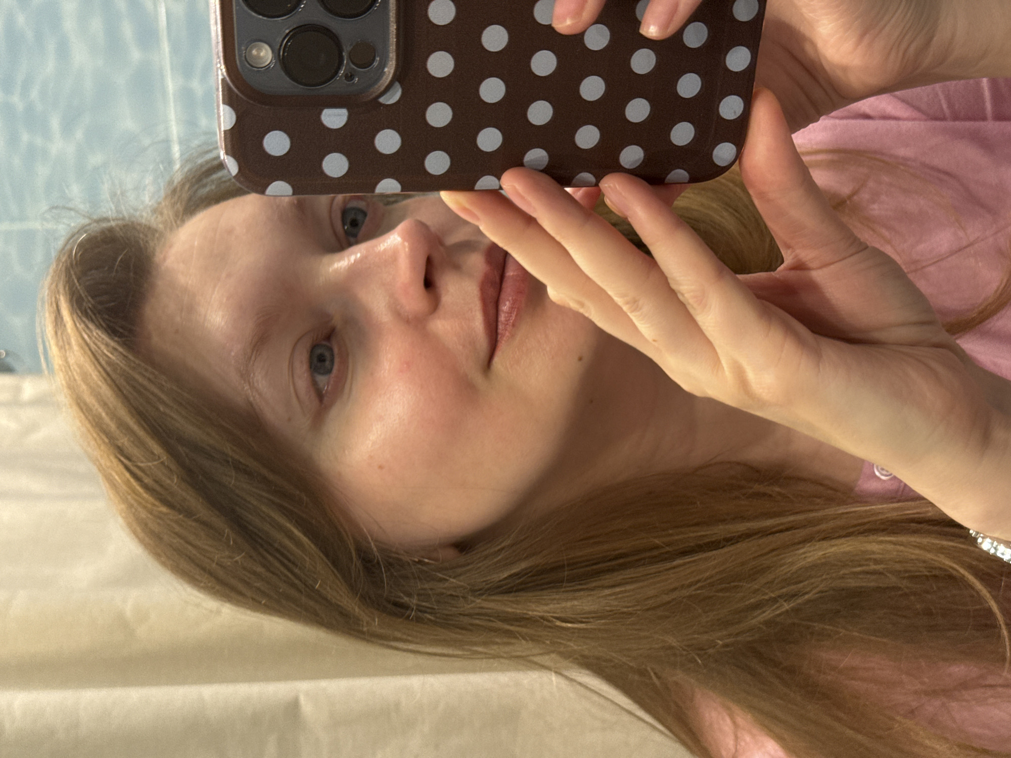 A fair-skinned person with light brown hair is holding a brown and white polka dot smartphone case. The person's face is visible, showing blue eyes and light pink lips. The person is wearing a pink top. The background includes a light-colored wall, possibly in a bathroom. The location is unknown; there is no information in the image to indicate it is in Izhevsk, Russia.
