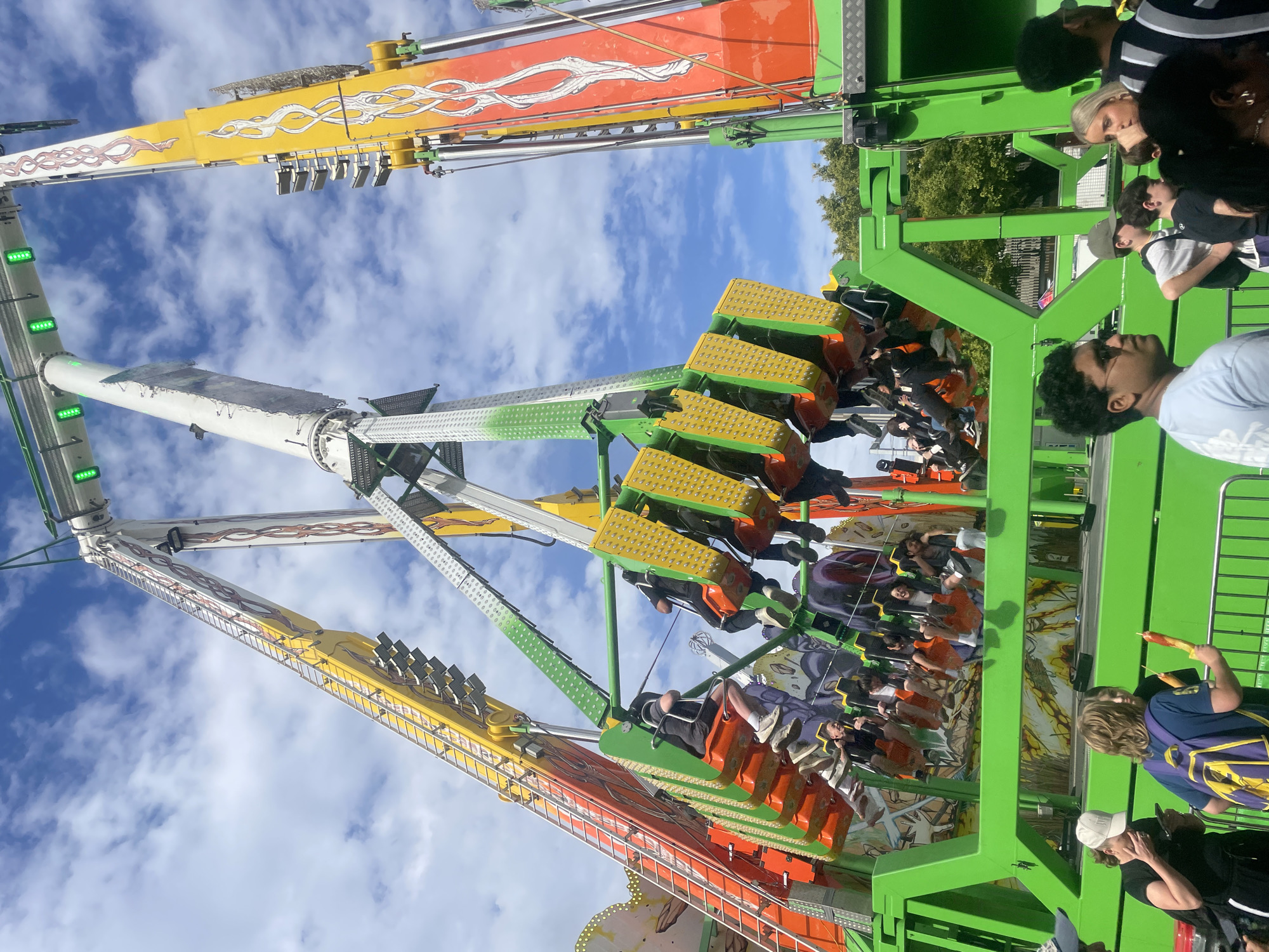 The image captures a thrilling amusement park ride, likely a pendulum ride, in operation at the Brisbane Ekka, a popular annual exhibition in Bowen Hills, Australia. The ride features large, brightly colored arms that swing riders in large arcs against a backdrop of a partly cloudy blue sky. Several people are visible, both on the ride in suspended seats and on the ground observing or waiting in line. The ride itself is a complex metal structure painted in vibrant green, yellow, and orange, adorned with flame-like decals. The surrounding environment suggests a bustling fairground, with glimpses of trees and other carnival attractions in the distance. The perspective is from ground level, looking up at the towering ride, emphasizing its scale and dynamic movement.