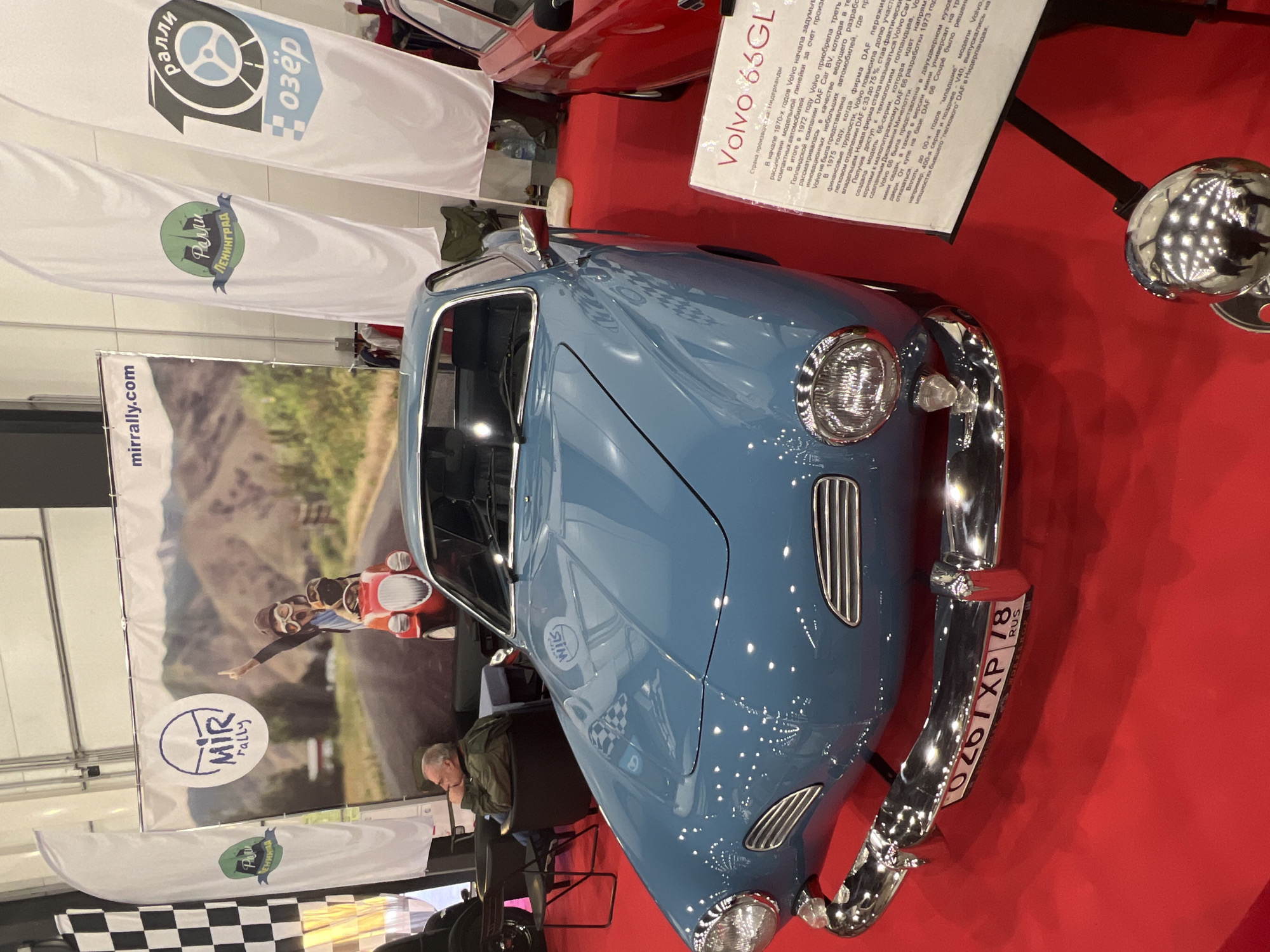 The image shows a blue Volvo 66GL car displayed on a red carpet. A sign next to the car provides information about its origins in the Netherlands. In the background, there's a large poster promoting "MIR rally" events, with a picture of a vintage car. The location appears to be an indoor exhibition hall, with flags bearing logos of "Ралли 10 Озёр" and "Ралли Ленинград" visible. An individual is seated in the background near the MIR rally poster.