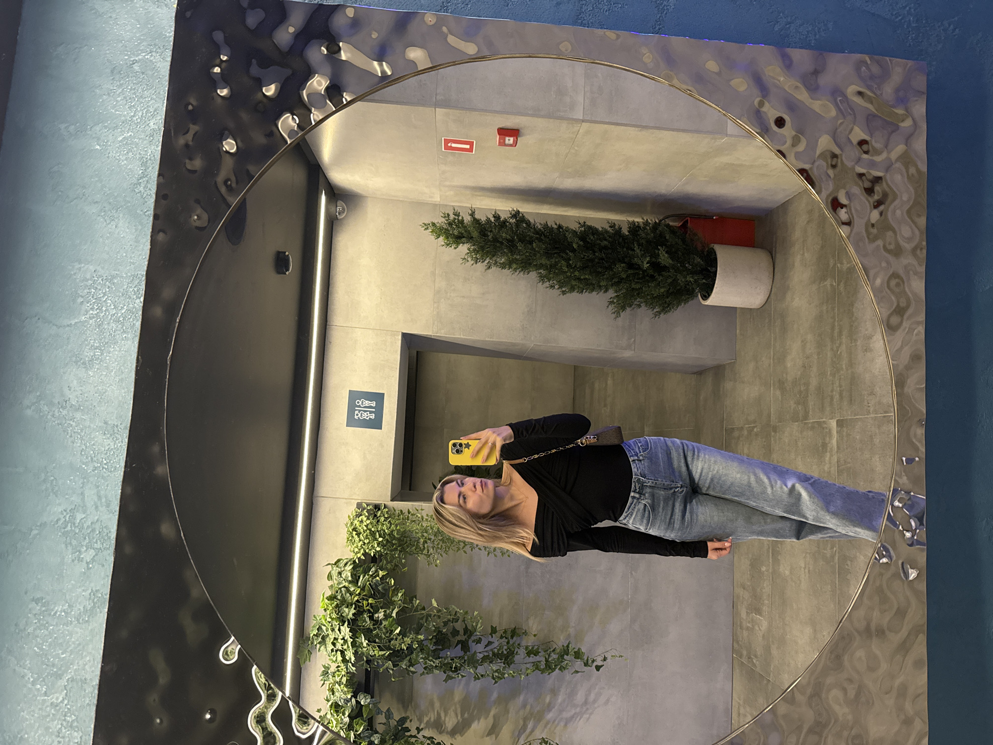 Here is a description of the image:
The image shows a woman taking a selfie in front of a large, round mirror with a decorative frame. The mirror reflects a hallway with gray tiled walls. There are two plants visible in the reflection, one potted in a white container and another growing vertically near the mirror's edge. The woman is wearing a black top and blue jeans, and she is holding a yellow smartphone. The setting appears to be a modern bathroom or public space with minimalist decor.